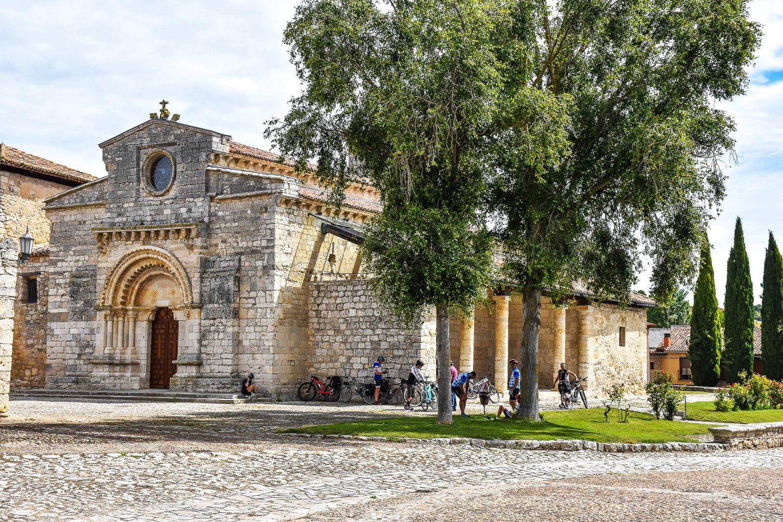 The stone Romanesque church of Santa María de la O in Wamba, with an arched doorway, a small round window and a roofed porch beside large trees