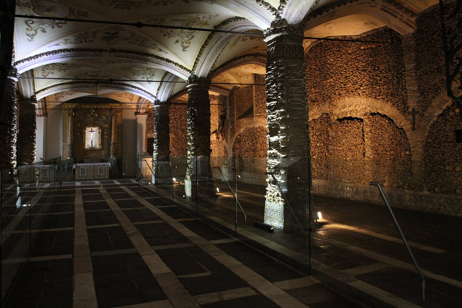 The nave of the Capela dos Ossos in Évora, with stone pillars and walls lined with bones, a vaulted ceiling and a lit altar at the far end