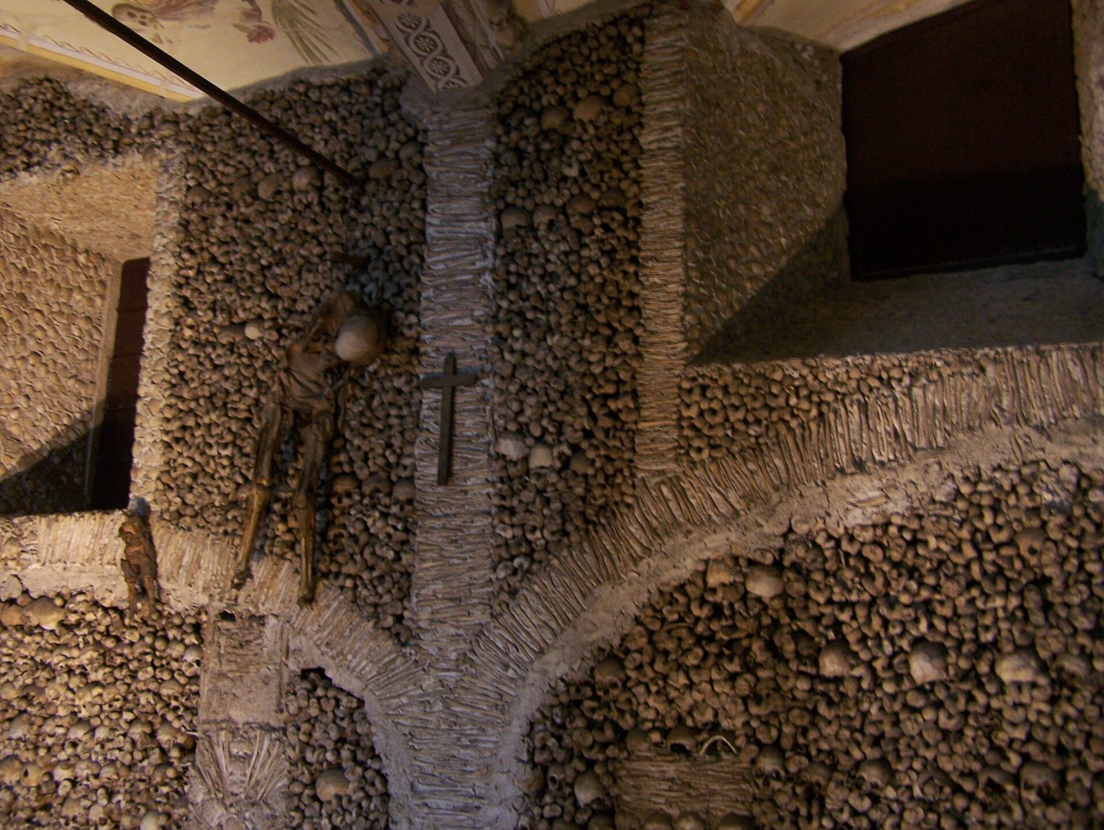 Walls and arches of the Capela dos Ossos in Évora covered in skulls and bones, with a small crucifix on a pier and a hanging figure to the left