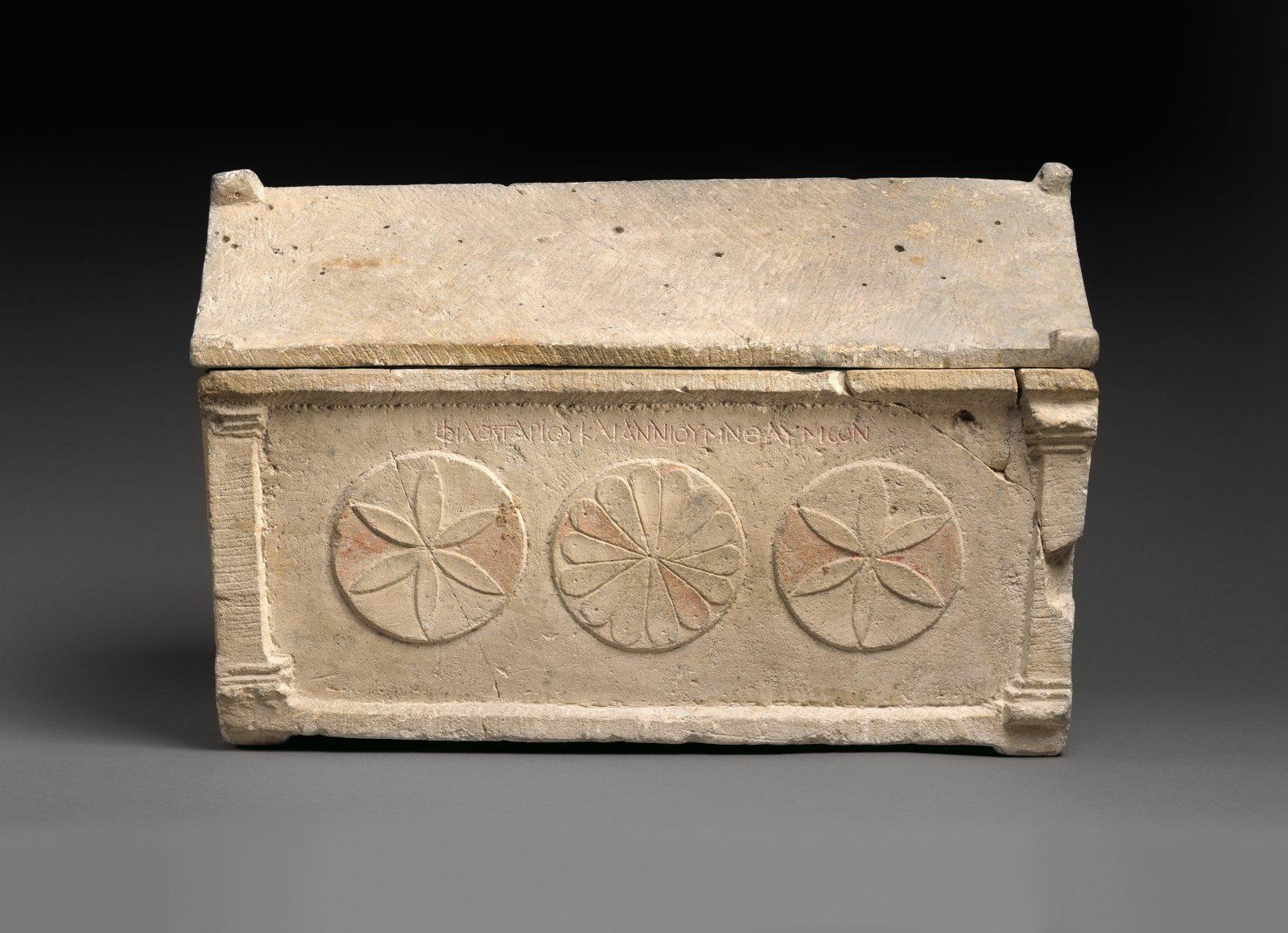 A small Roman limestone ossuary with a gabled lid and three carved rosettes on the front, with a line of inscribed lettering above them