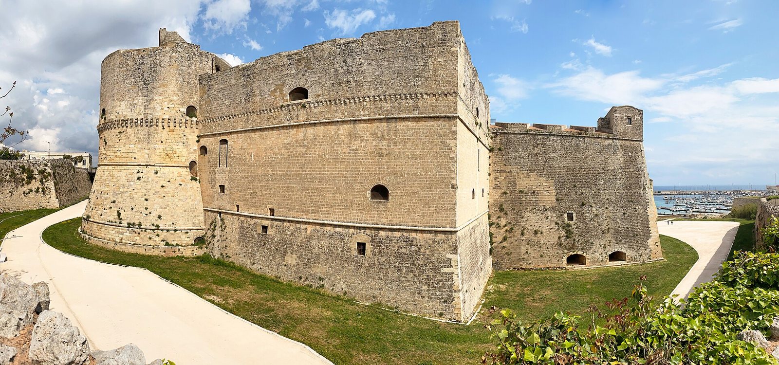 The Aragonese castle of Otranto, a stone fortress with a round tower on the left and a bastion on the right, over a green lawn and a path