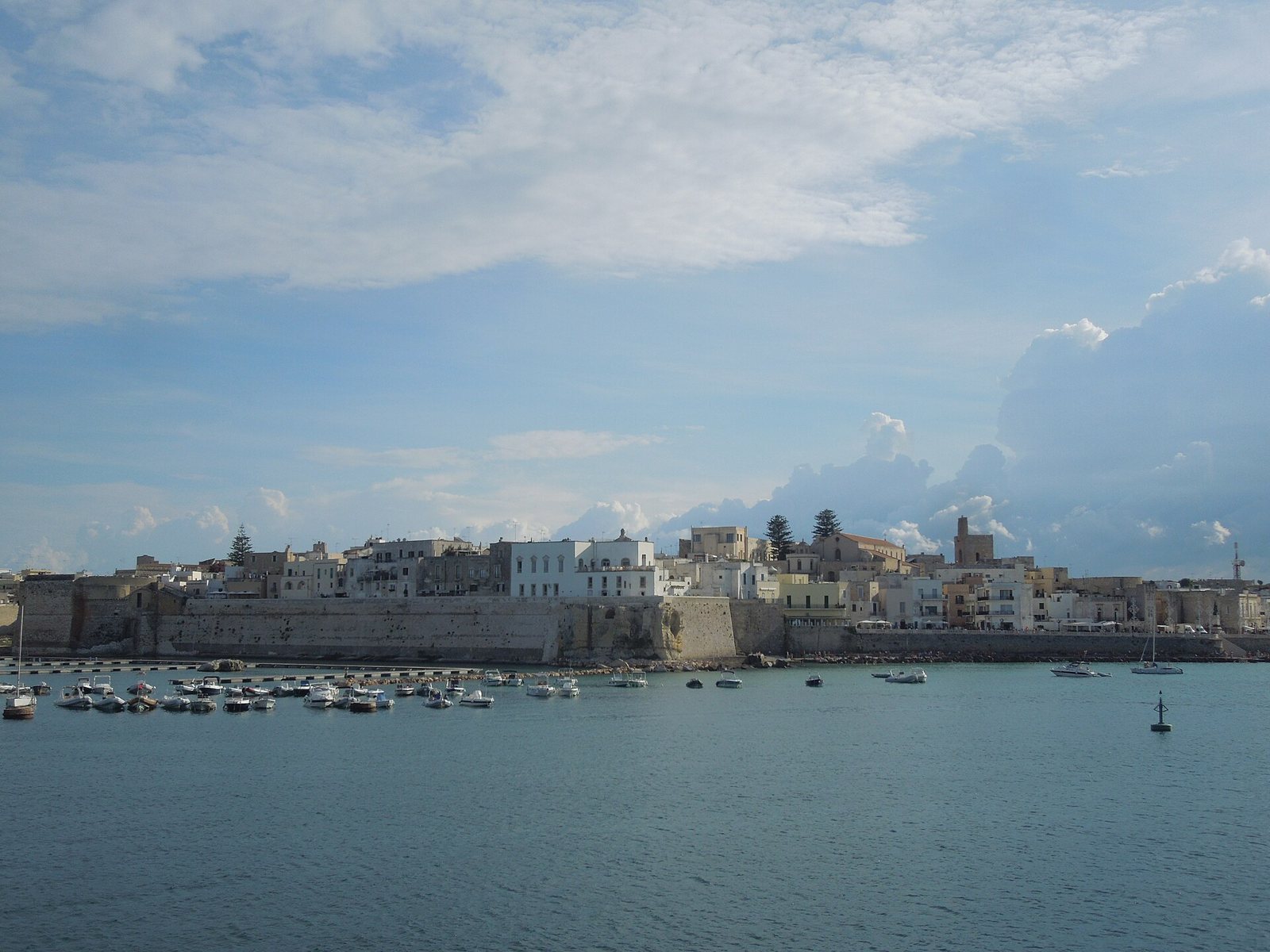 The old town and harbour of Otranto seen from the water, with white and ochre houses behind a sea wall, small boats and a cloudy blue sky
