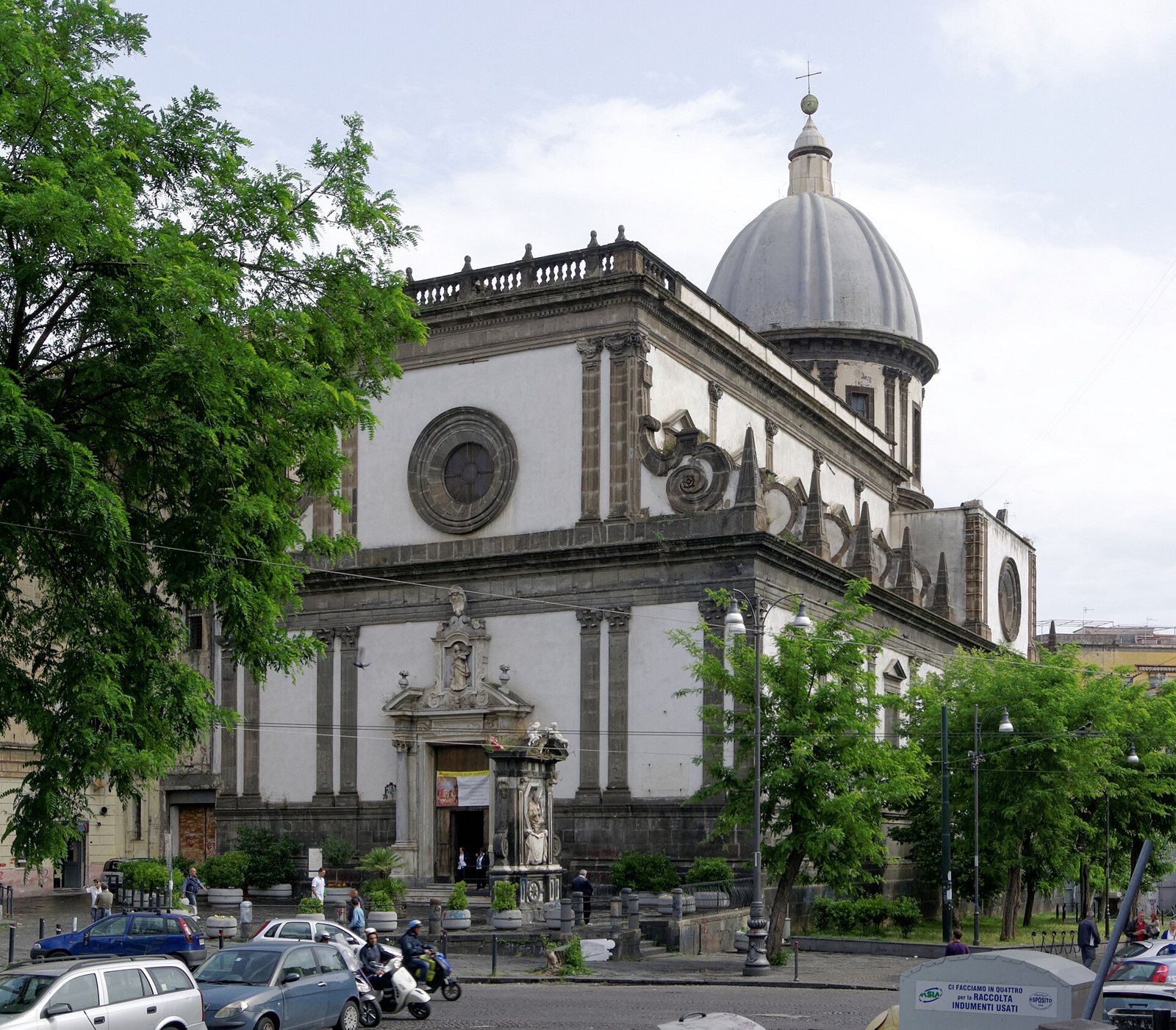 The front of the church of Santa Caterina a Formiello in Naples, with a pale dome, round windows and a street with parked cars in front