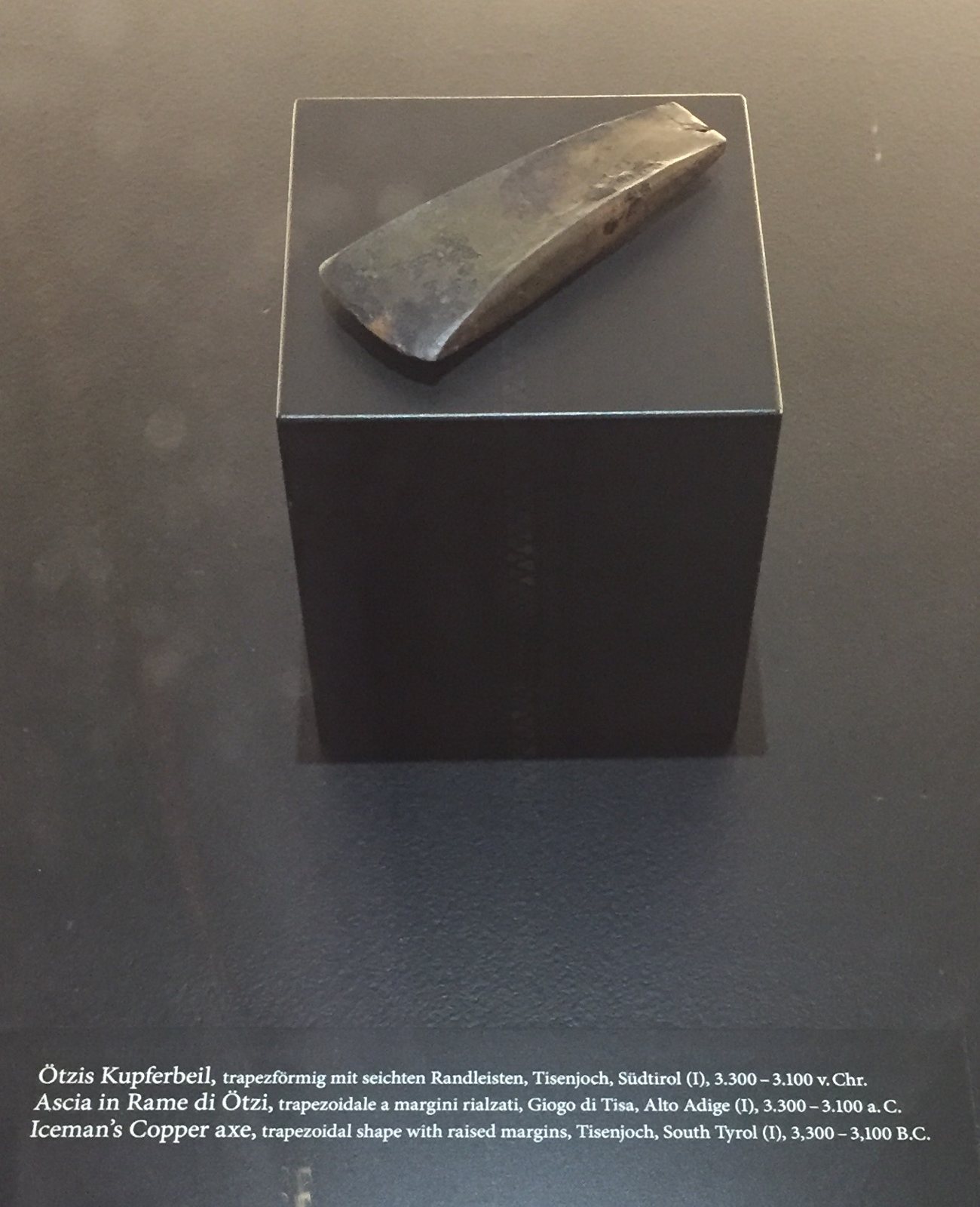 A flat, trapezoid copper axe blade resting on a dark display block in a case, with a museum label below it