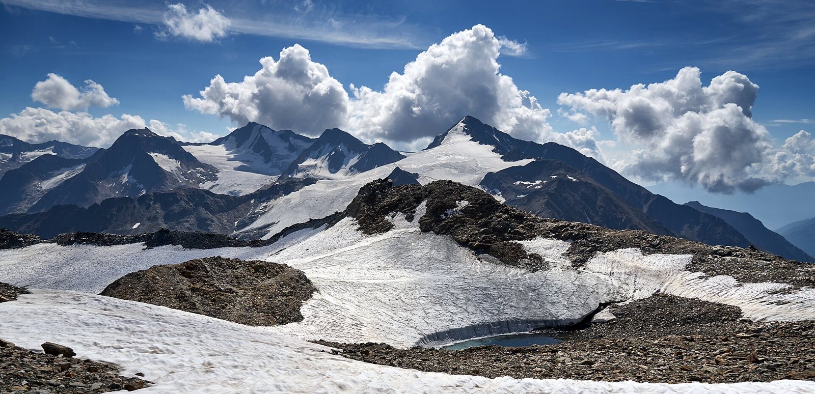 Snowfields and a small meltwater pool in the foreground below the rocky, snow-streaked peaks of the Similaun and Schnals ridge in the Ötztal Alps, under towering white clouds