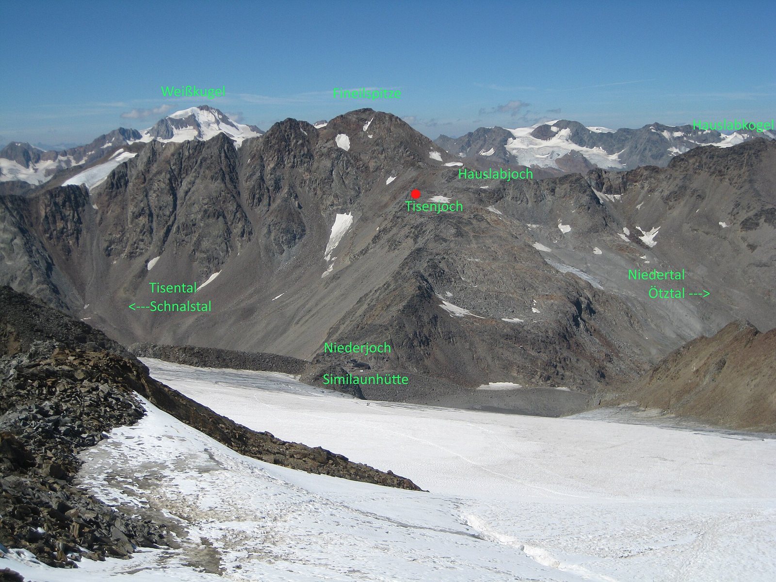 A view across a snowfield to a rocky mountain ridge in the Ötztal Alps, with green labels naming the peaks and passes and a red dot marking the find site at Tisenjoch