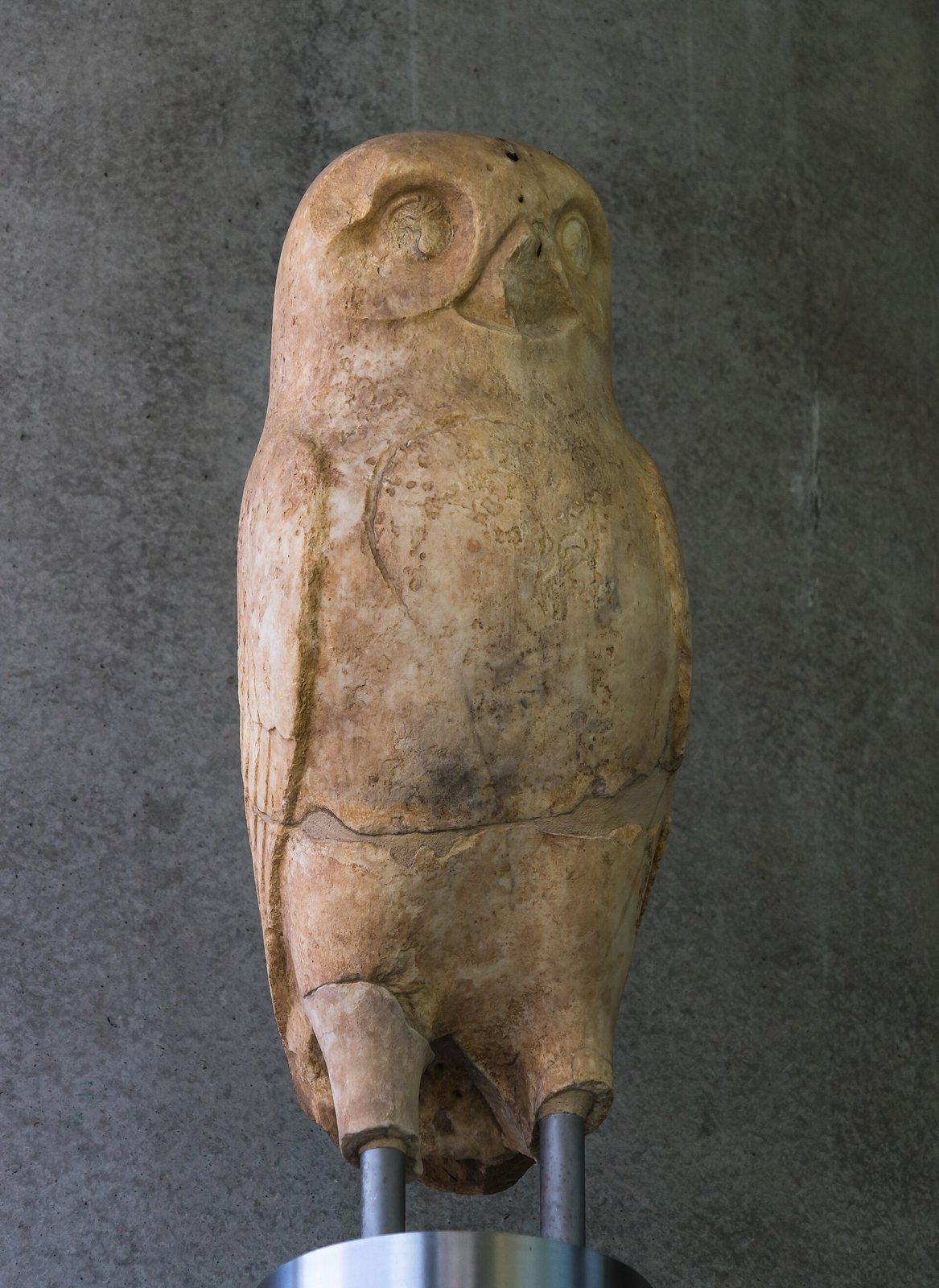 An ancient stone owl with large round eyes, its feet missing, mounted on metal rods in the Acropolis Museum