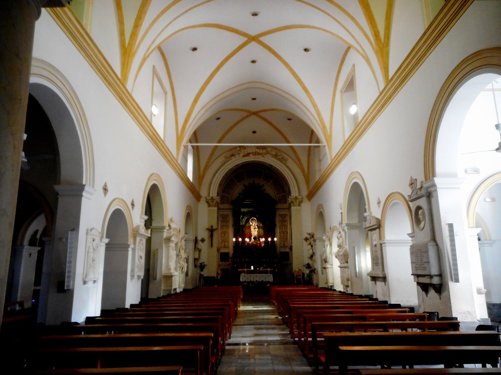 The nave of the Capuchin church in Palermo, with a pale vaulted ceiling picked out in gold, rows of wooden pews and a candlelit altar at the far end