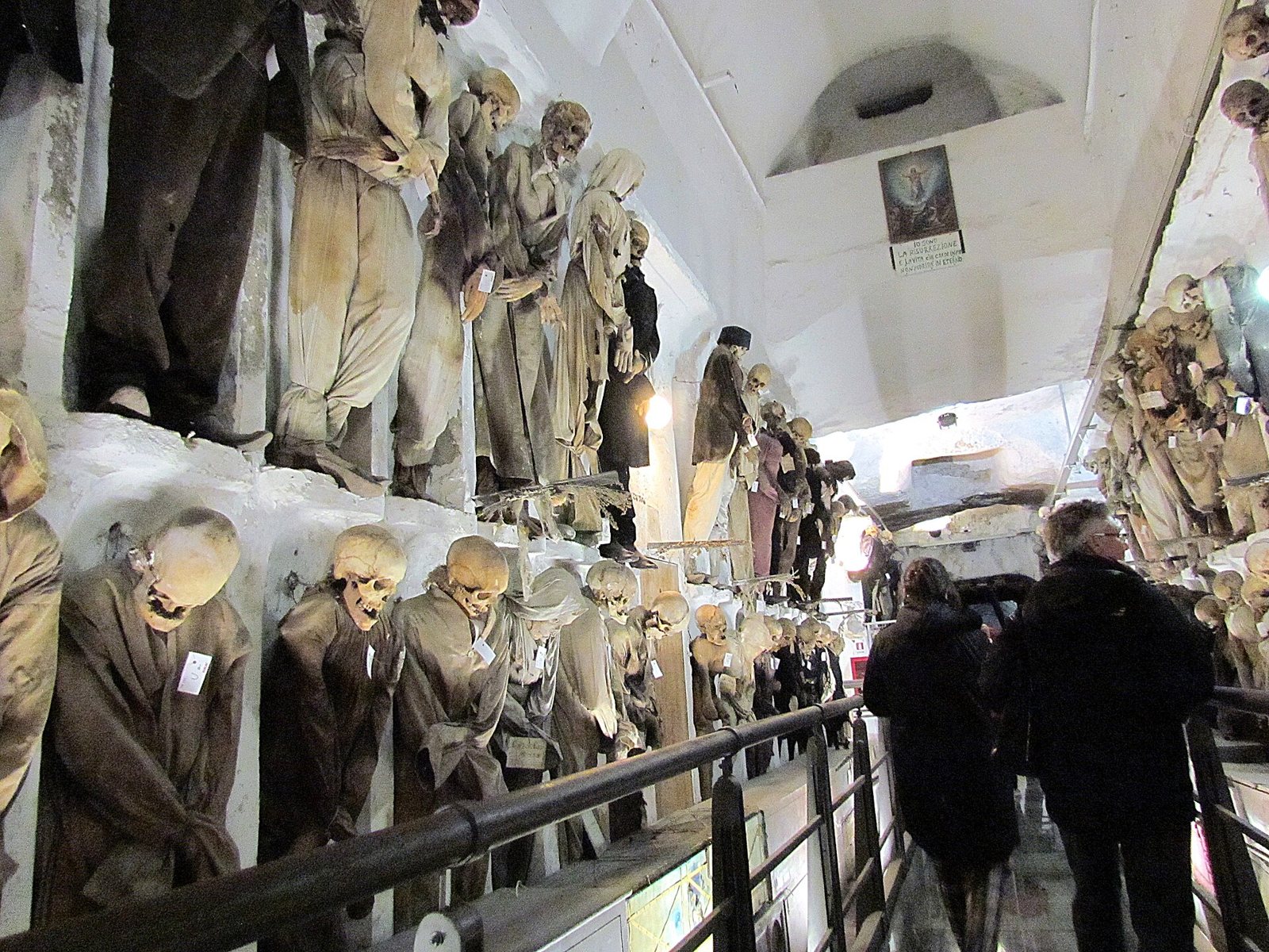 A railed walkway in the Capuchin Catacombs, with rows of clothed mummified bodies standing on ledges along the wall and two visitors walking away from the camera
