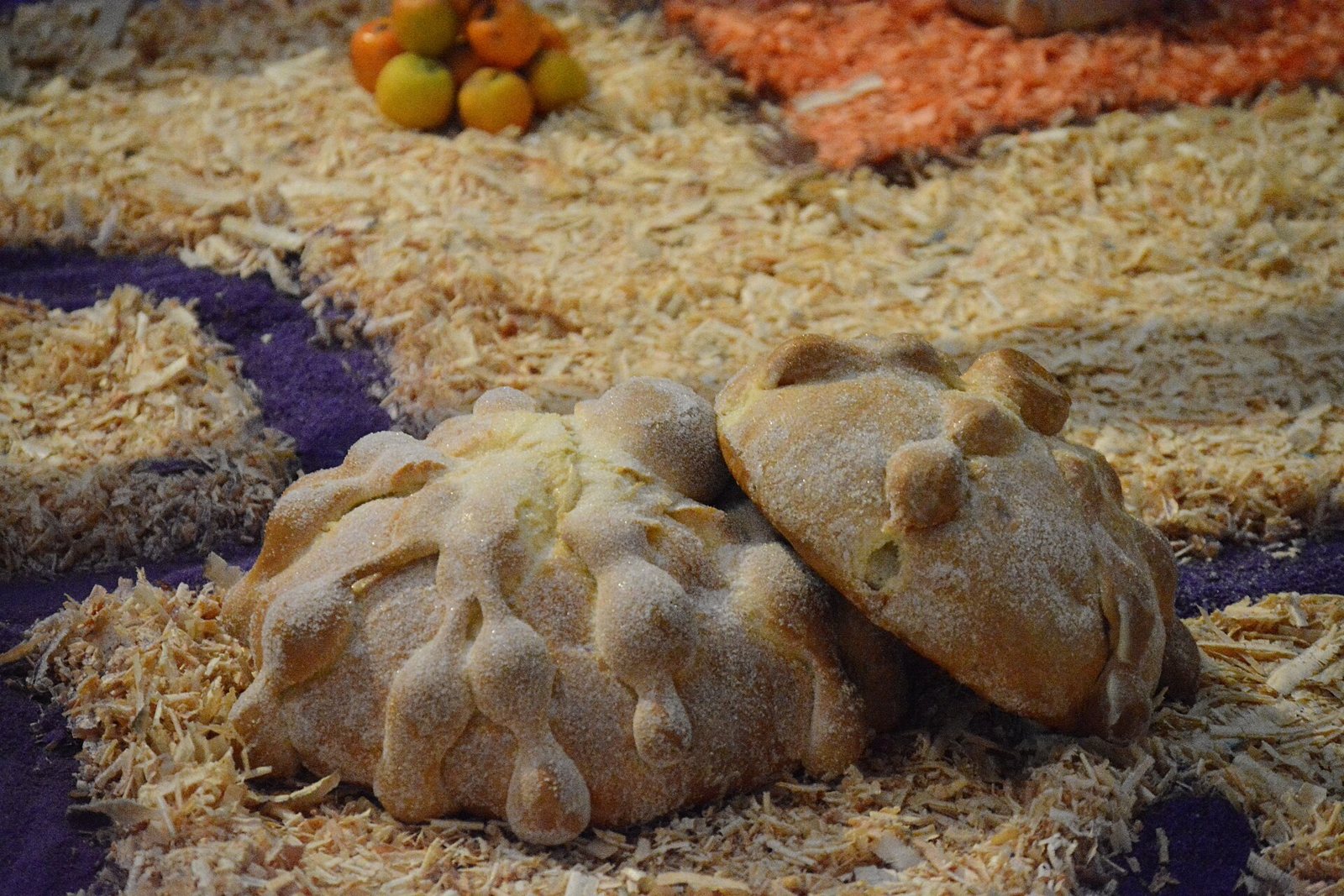 Two round pan de muerto loaves decorated with dough bones resting on a bed of wood shavings on an altar, with tangerines in the background