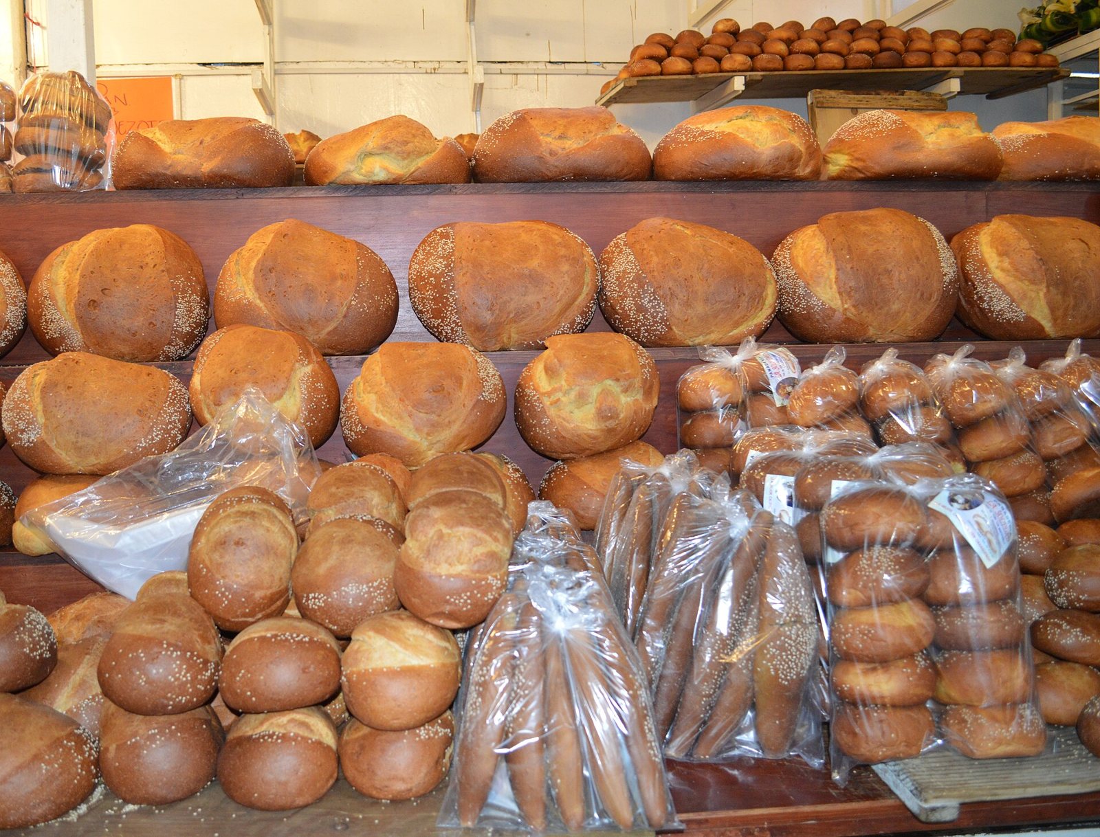 Shelves of round, sesame-topped pan de yema loaves stacked in a market stall, with smaller rolls in plastic bags below