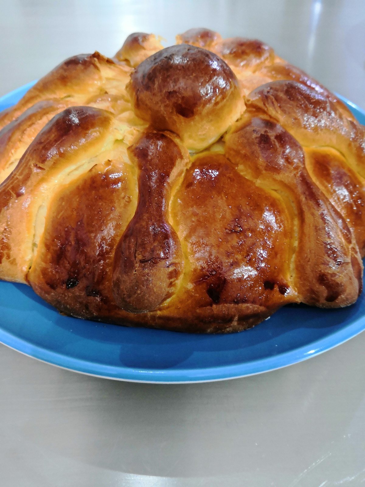 A glossy round homemade pan de muerto on a blue plate, with bone-shaped strips running down its sides and a round knob on top