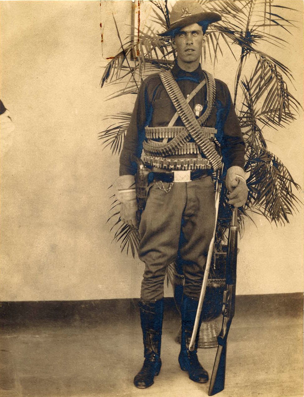 Sepia studio portrait of Emil Holmdahl standing in uniform with crossed bandoliers and a sabre, in front of potted palms