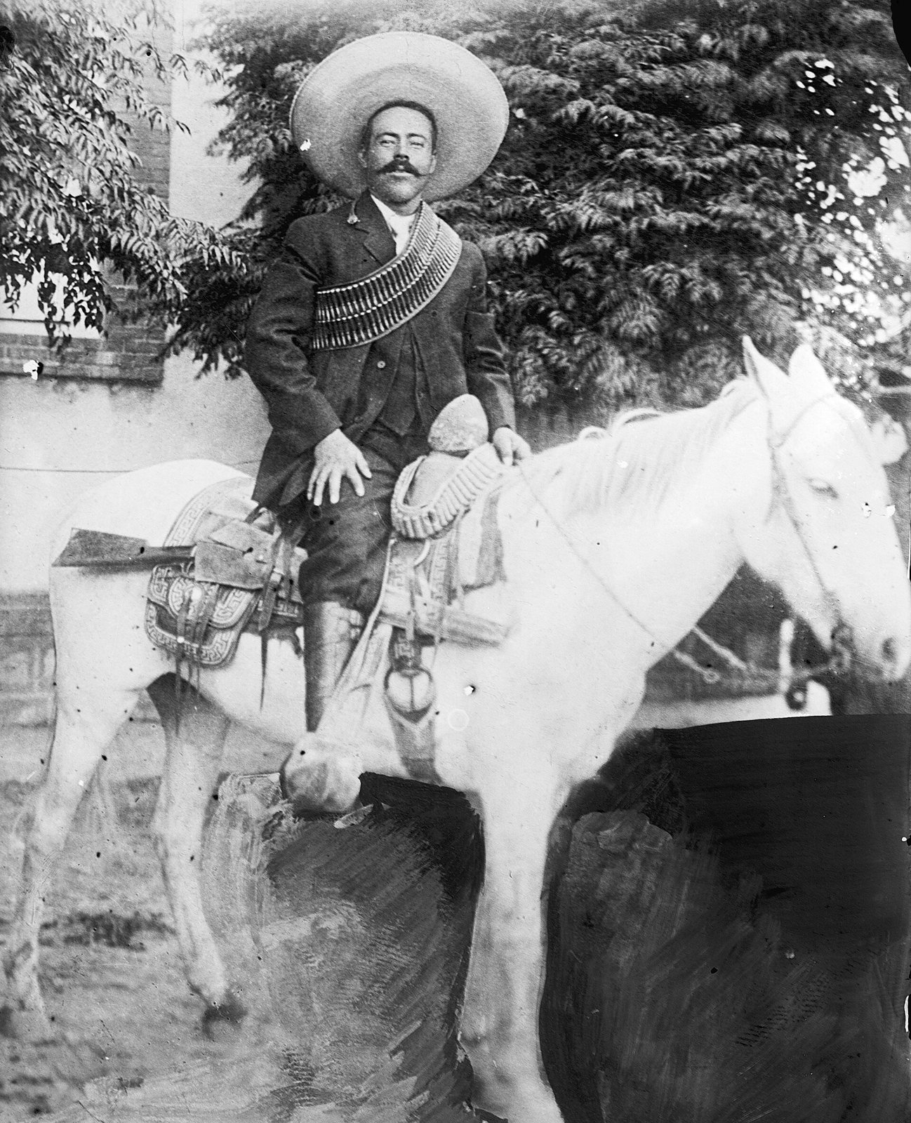 Black-and-white photograph of Pancho Villa in a wide sombrero and bandolier, seated sideways on a pale horse in front of trees