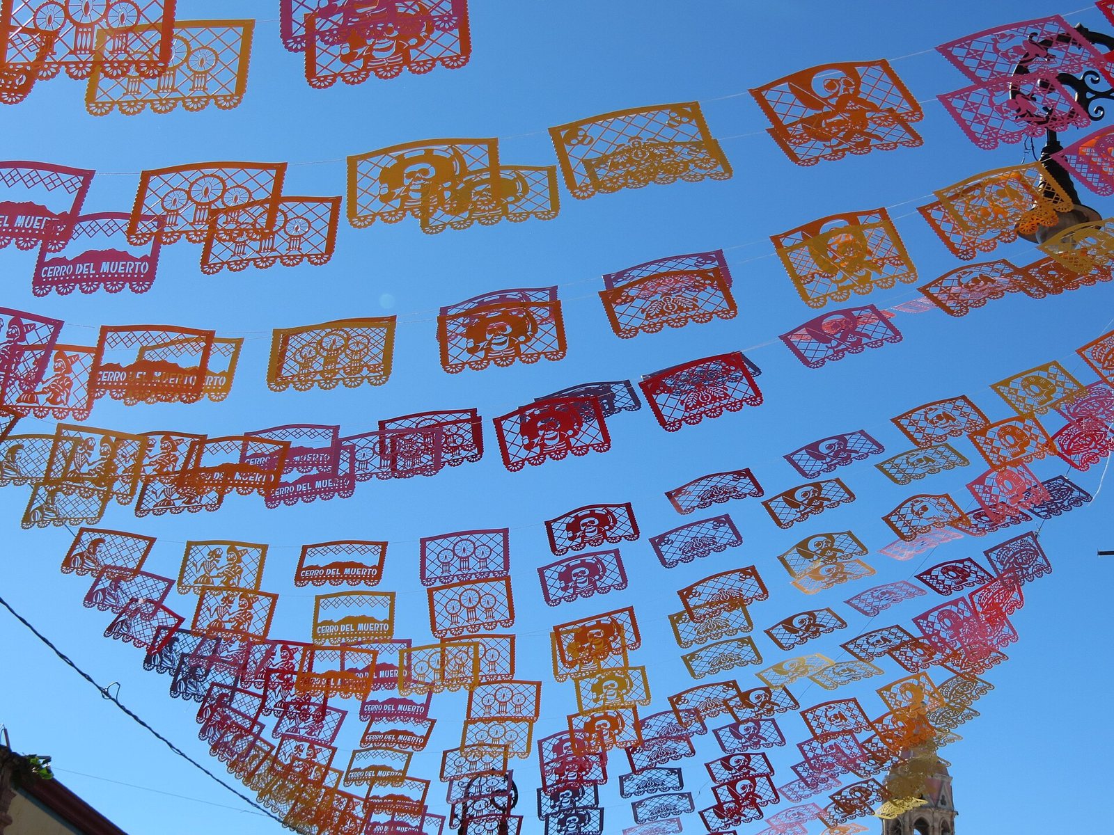 Rows of orange, pink and red cut-paper banners with skulls, catrinas, candles and the words Cerro del Muerto strung across a blue sky