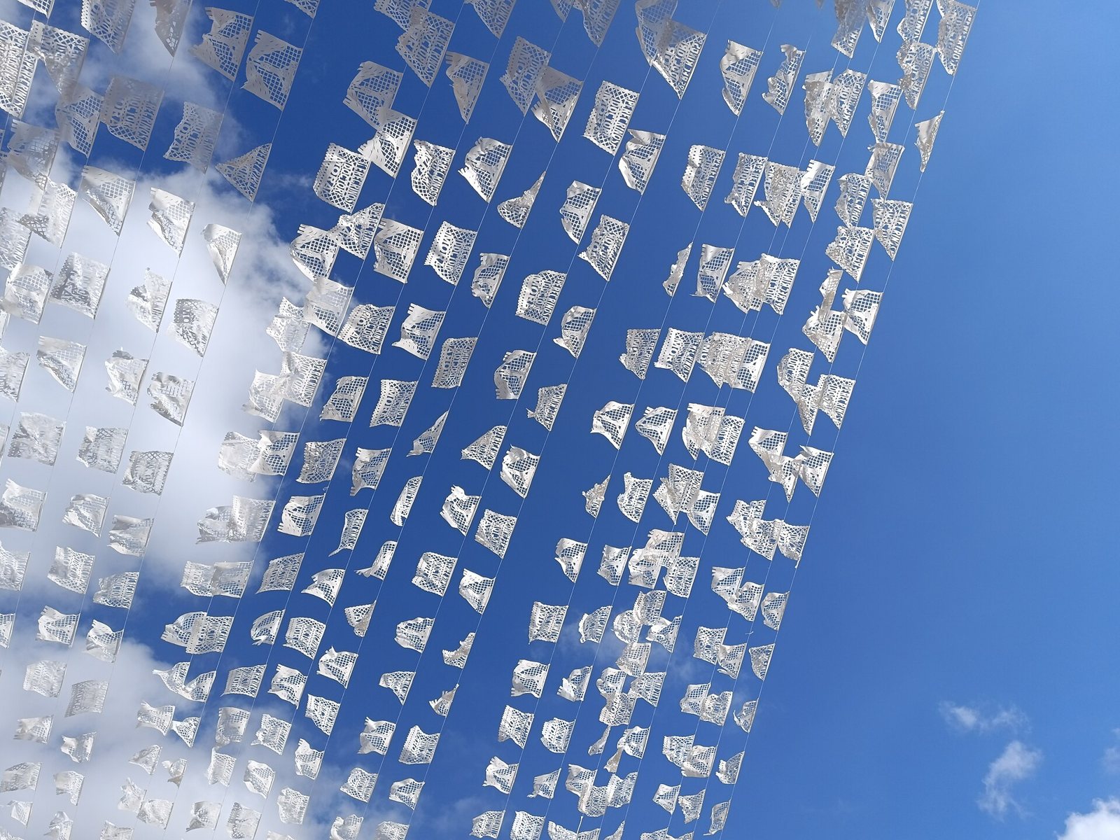 Long strings of small white papel picado flags with lattice cut-outs fluttering against a blue sky and white clouds