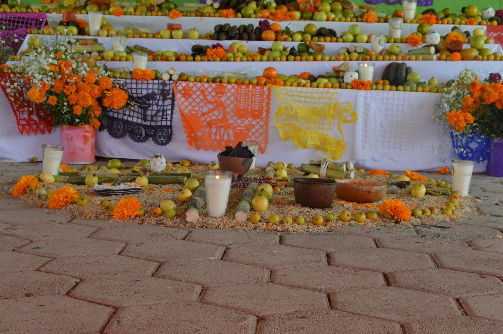 Cut-paper banners in purple, orange, yellow and white, with cut-out skeletons and skulls, hung along a tiered ofrenda laden with fruit, sugar cane, candles in glasses and cempasúchil, above a floor arrangement of fruit and clay bowls
