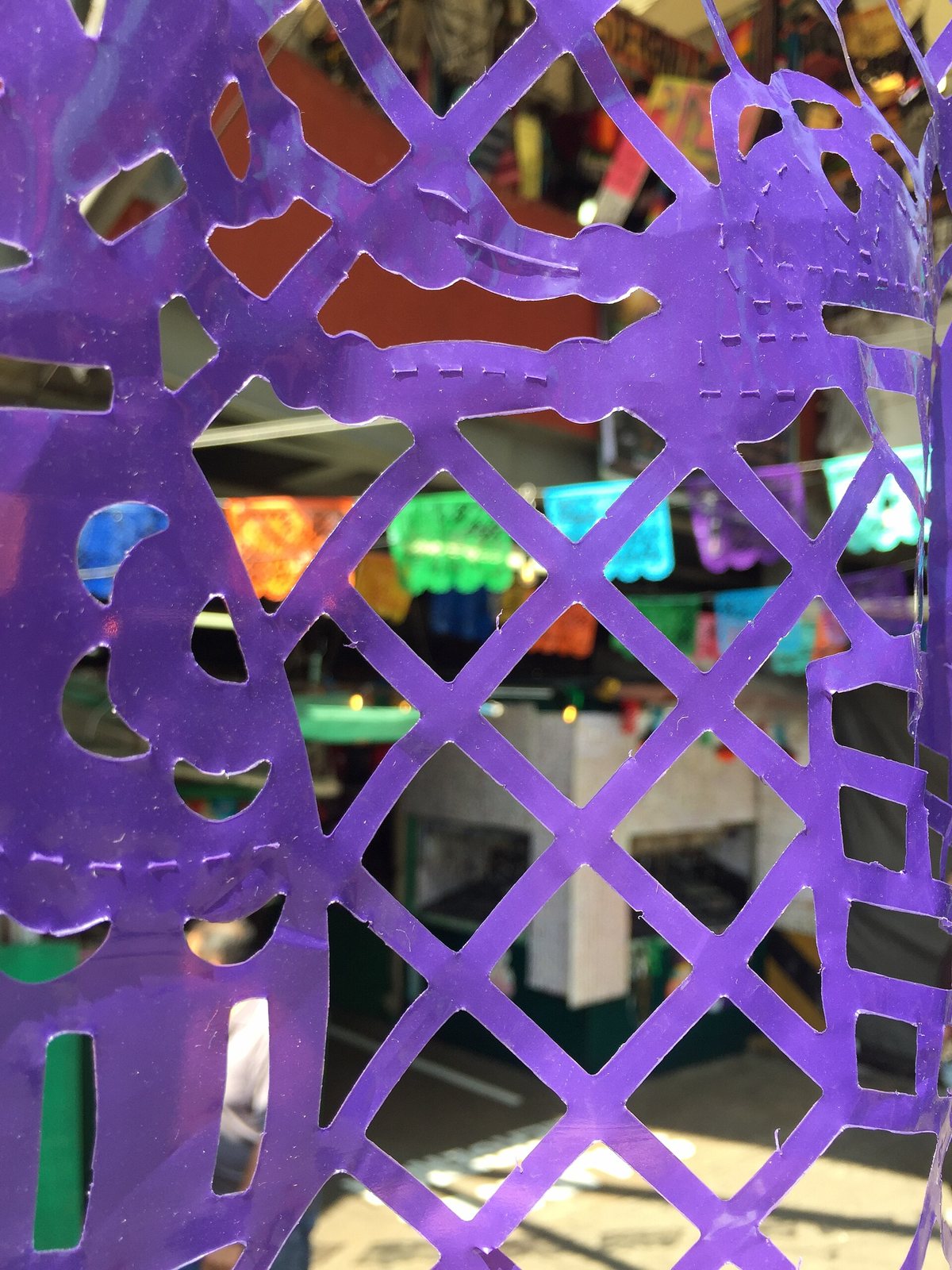 Close view of a purple plastic papel picado sheet cut in a diamond lattice, with strings of colored banners hanging in a market behind it