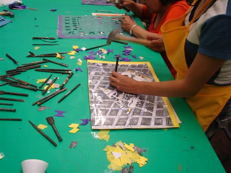 Hands striking a chisel with a hammer through a stack of paper under a printed skeleton pattern, on a green table scattered with chisels and purple and yellow paper scraps