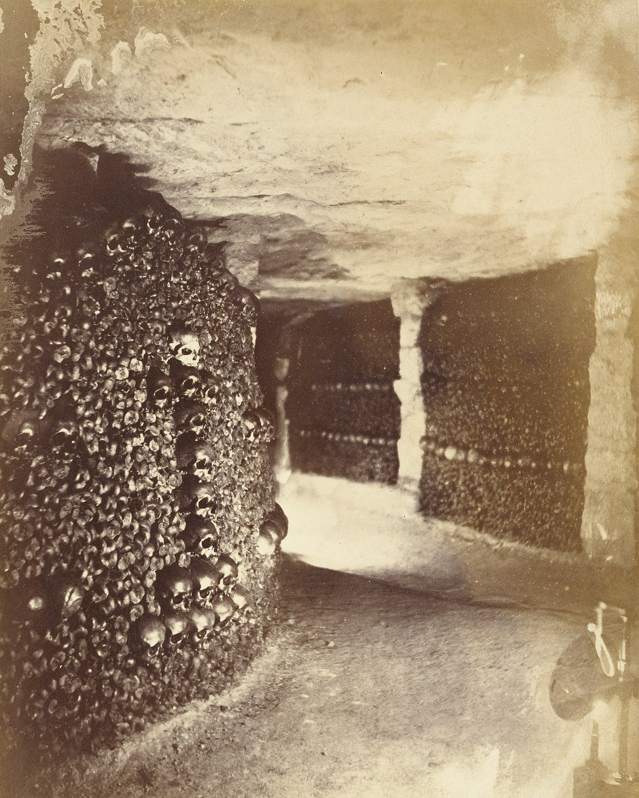 A sepia photograph of a catacomb gallery with walls of stacked skulls and bones and a pillar, taken with artificial light