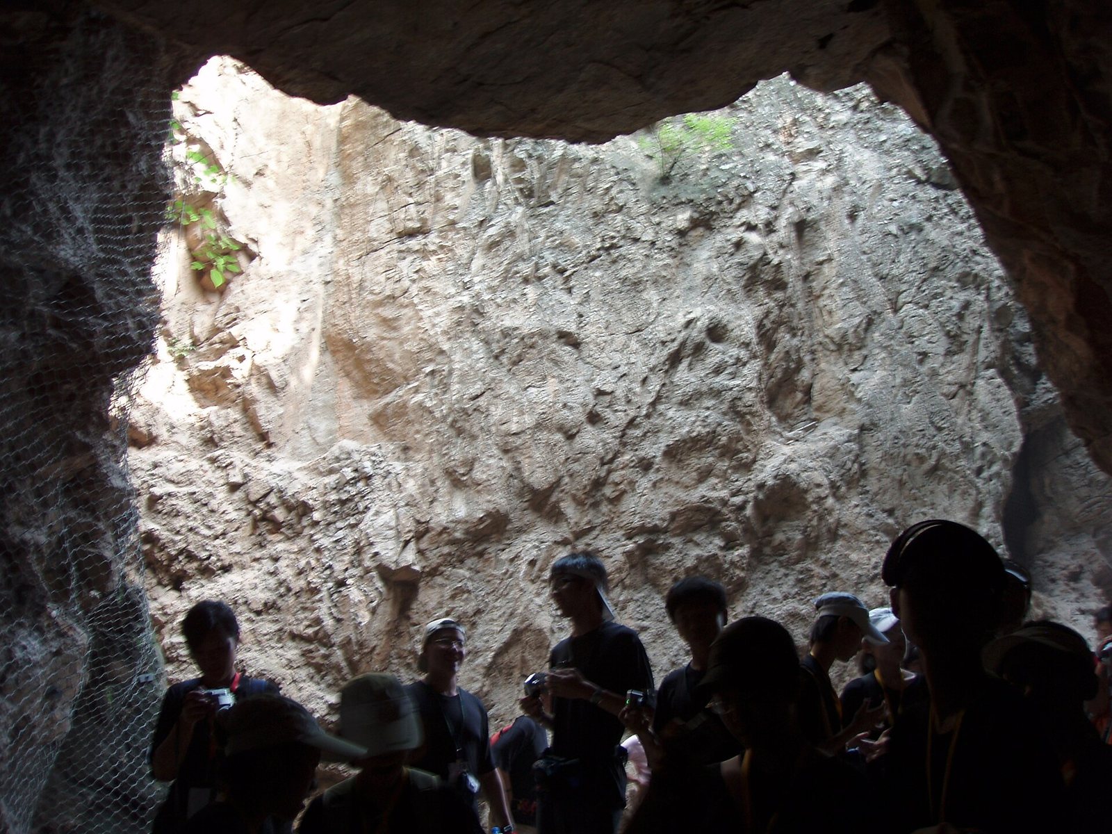 Looking up from inside a Zhoukoudian cave at a high rock wall and an opening to daylight, with the dark silhouettes of visitors at the lower right