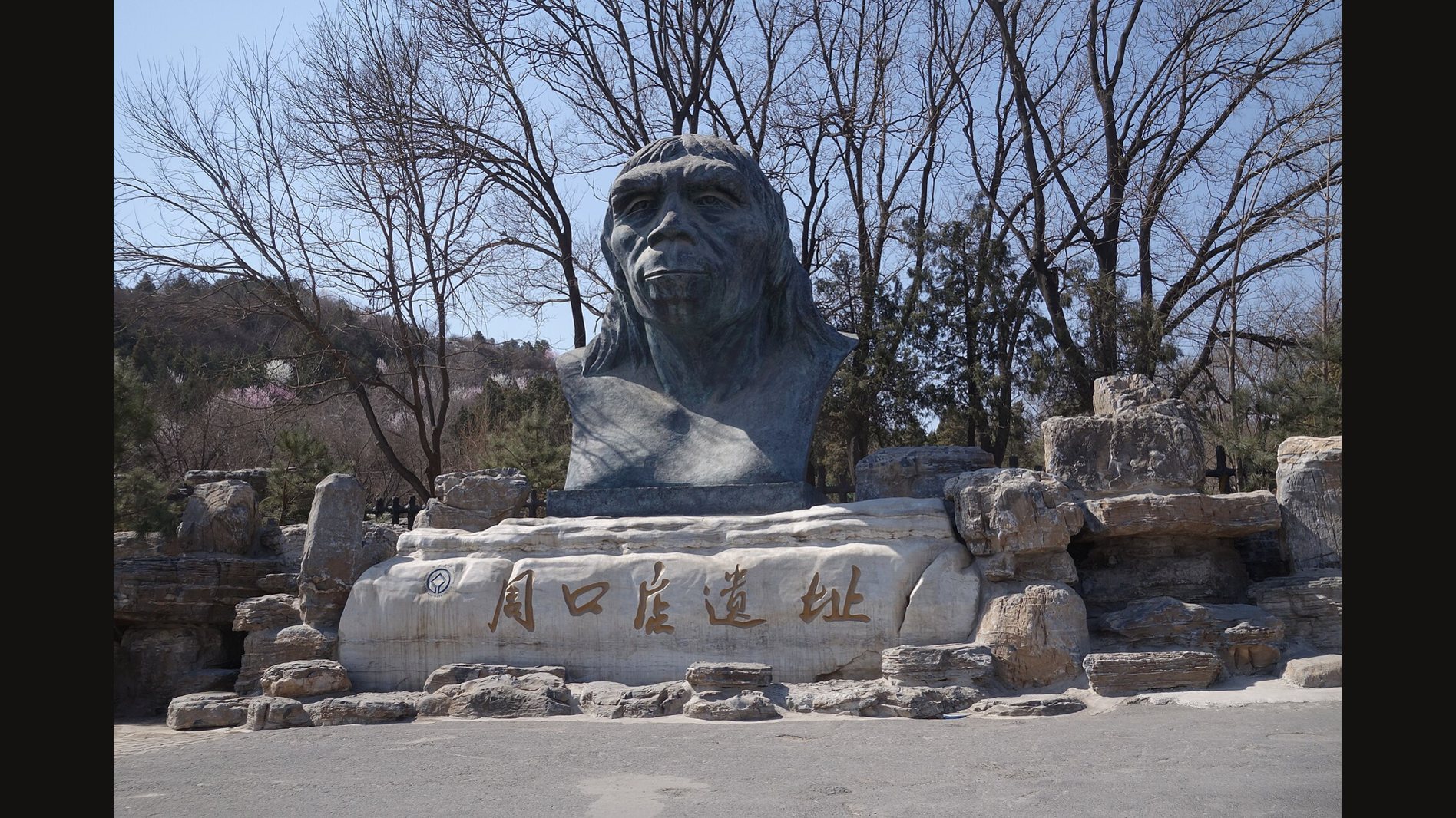 Dark stone sculpture of a Peking Man head above a carved stone base with Chinese characters, among rocks and bare trees at the entrance to the Zhoukoudian site