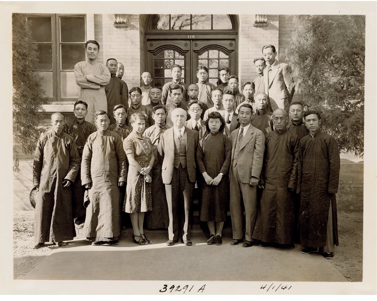Sepia group photograph of about thirty scientists and staff standing in front of a building doorway, men in long gowns and suits and two women in the front rows, with a date in handwriting at the bottom