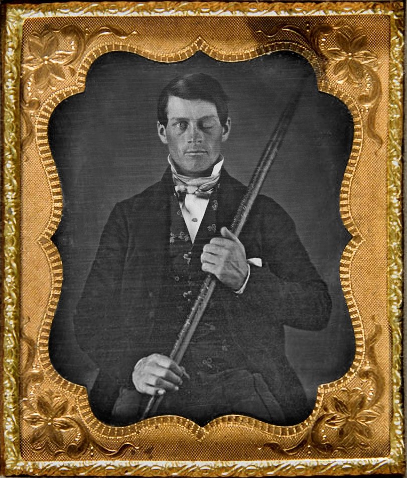A cased daguerreotype in an embossed gold mat: a young man in a dark coat and patterned vest, his left eye closed, holding a long tamping iron diagonally across his body