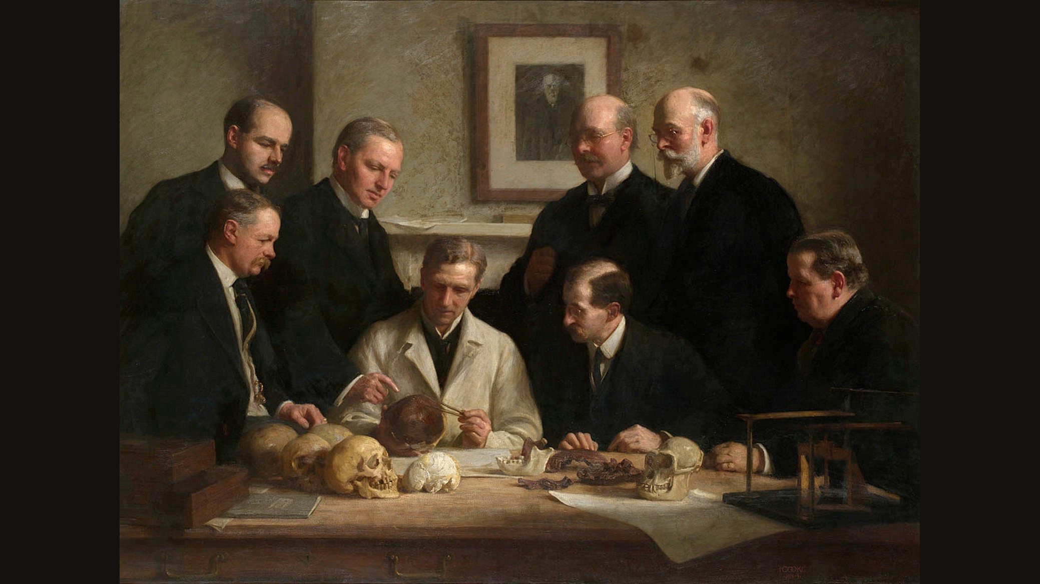 Oil painting of eight men in dark suits gathered around a table of skulls and bones, one in a white coat holding a skull piece, with a portrait on the wall behind them