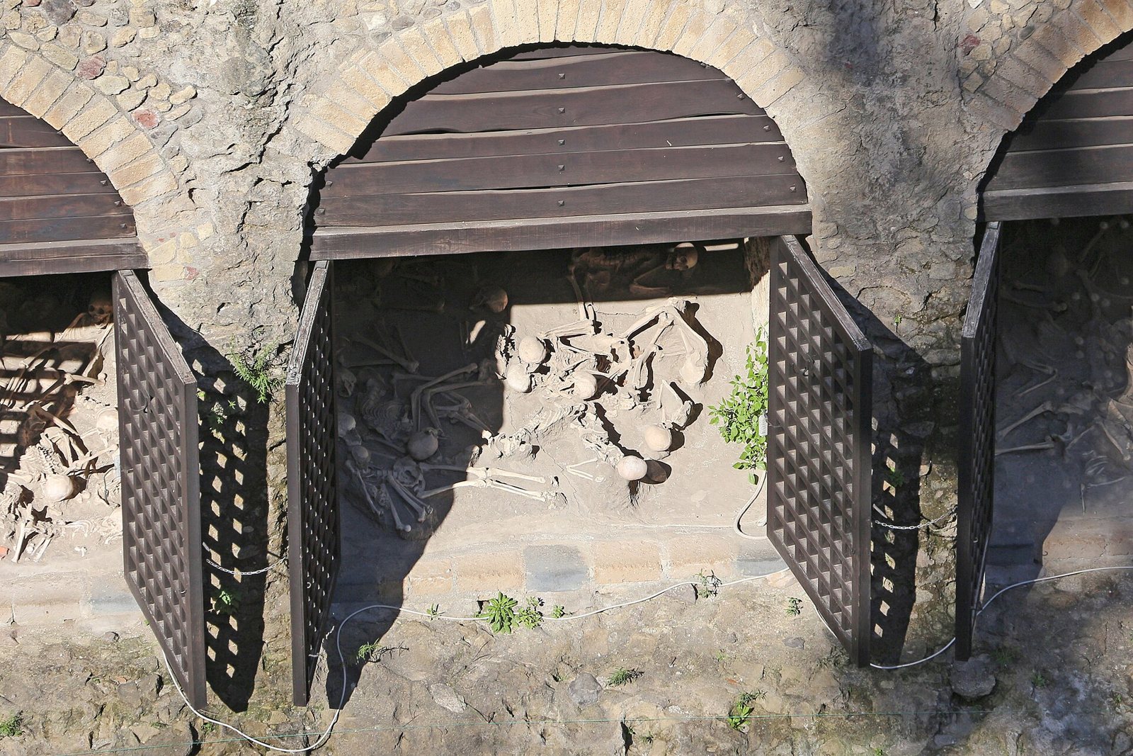 Vaulted stone arches of the boat chambers at Herculaneum, with open dark lattice doors and wooden hoods, and human skeletons visible on the ground inside