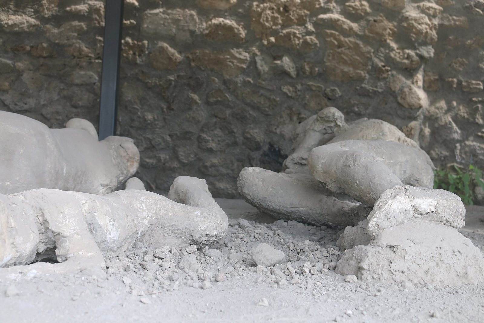 Pale plaster casts of two figures lying on grey gravel in front of a rough stone wall, in a covered outdoor display at Pompeii