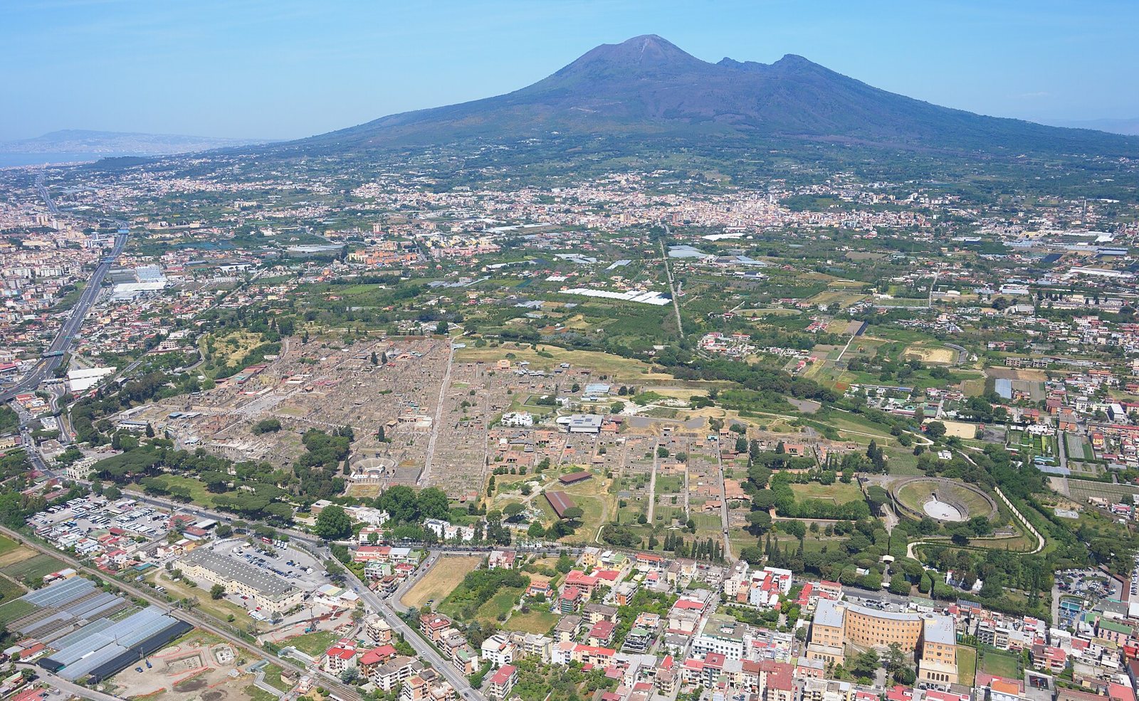 Aerial view of the excavated ruins of Pompeii among green fields and towns, with Mount Vesuvius rising behind under a blue sky