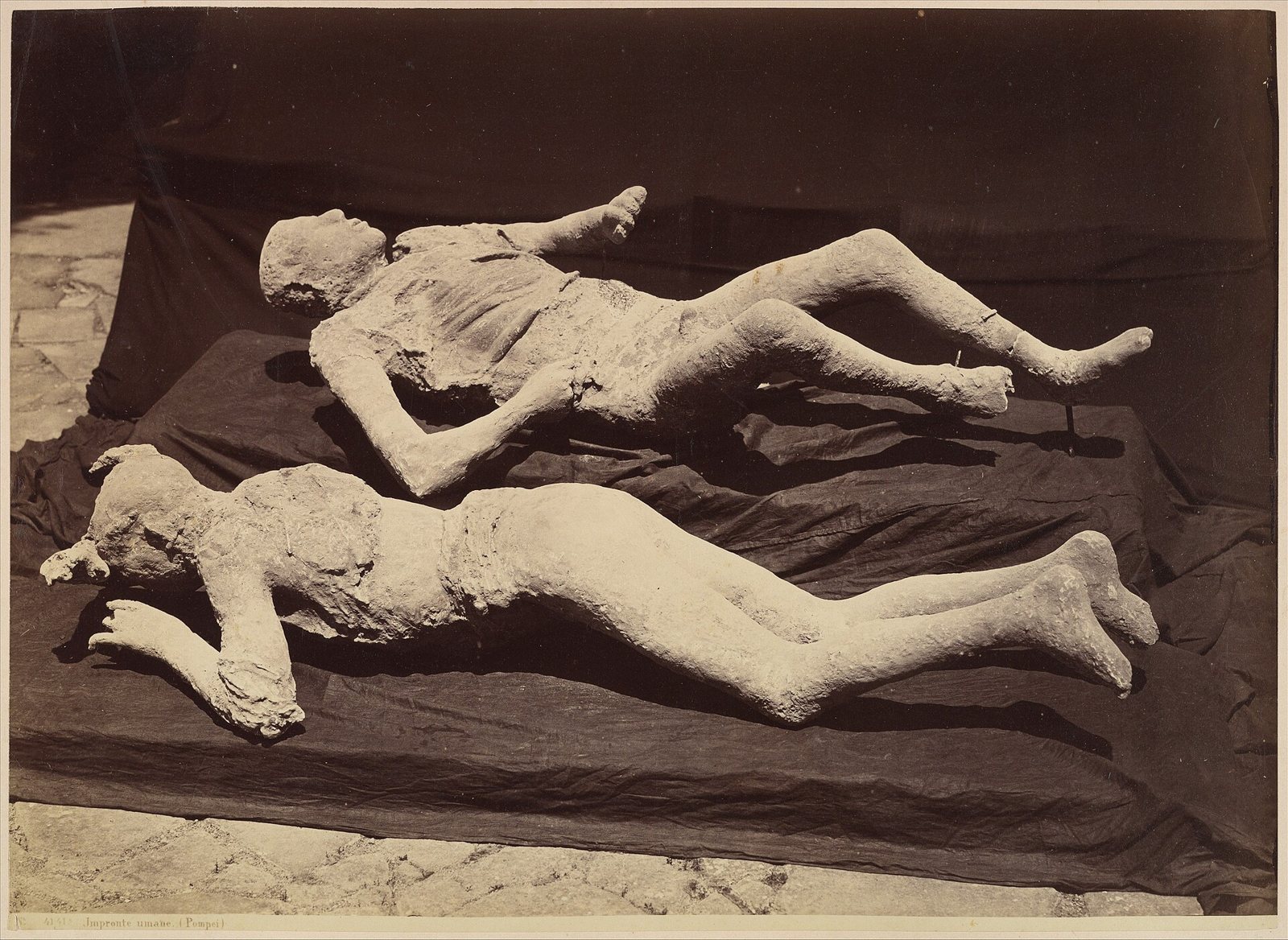 Sepia photograph of two plaster casts of bodies lying side by side on dark cloth, one on its back and one face down, with the folds of clothing visible