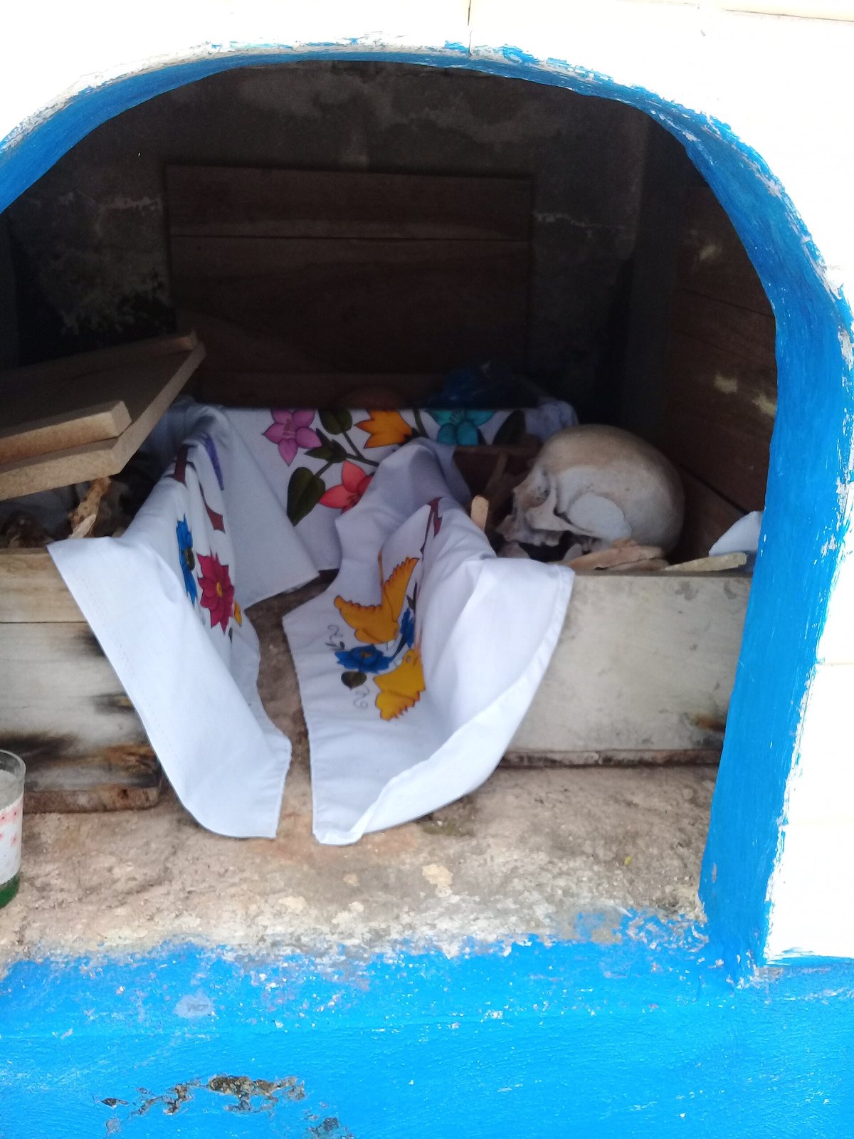 A small whitewashed niche painted blue at the edges, holding a skull and bones on a white cloth embroidered with colorful flowers and birds