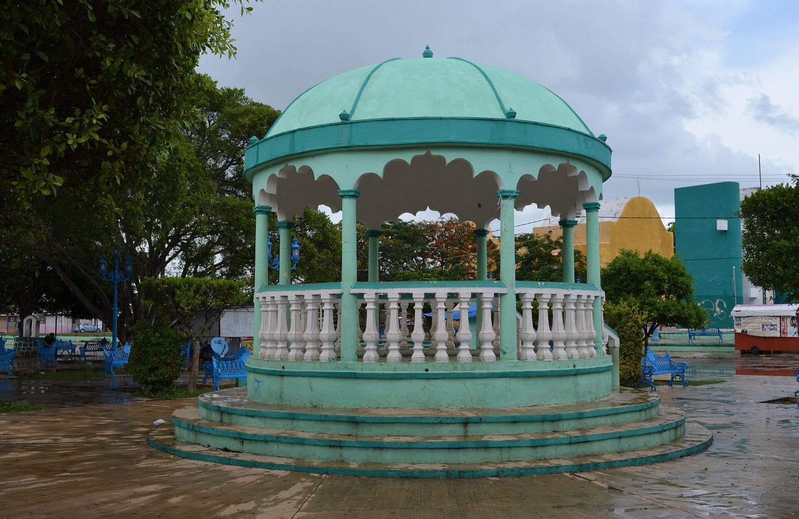 The main plaza of Pomuch after rain: a round turquoise bandstand with a white balustrade among trees, and a yellow building behind it