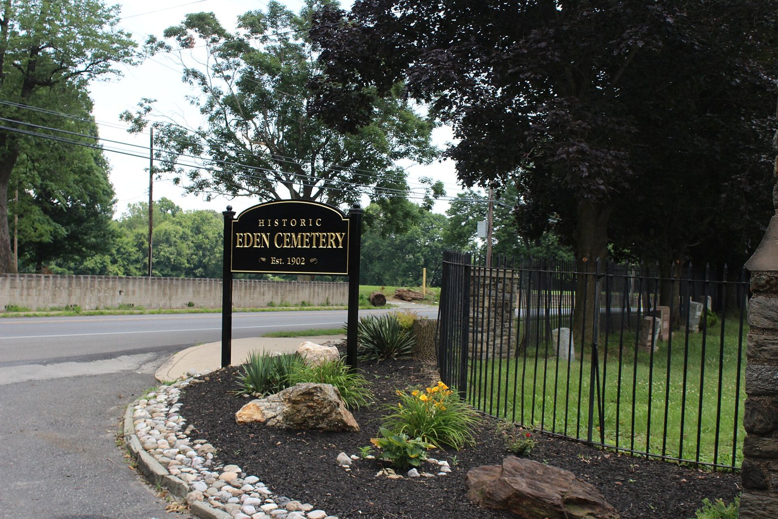 Un letrero negro con letras doradas que dice Historic Eden Cemetery, Est. 1902, junto a una reja de hierro y un parterre bordeado de piedras, bajo árboles altos, en la entrada del cementerio junto a la carretera