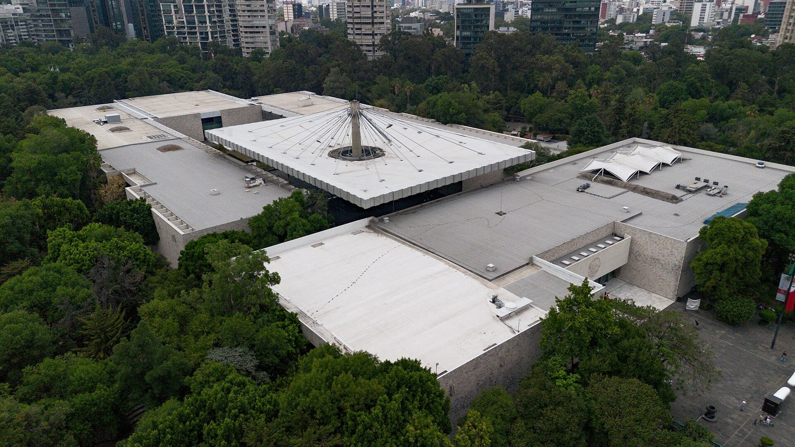 Vista aérea de los techos planos del Museo Nacional de Antropología de la Ciudad de México y su gran techo en forma de paraguas sobre el patio central, rodeado de árboles verdes con edificios de la ciudad al fondo