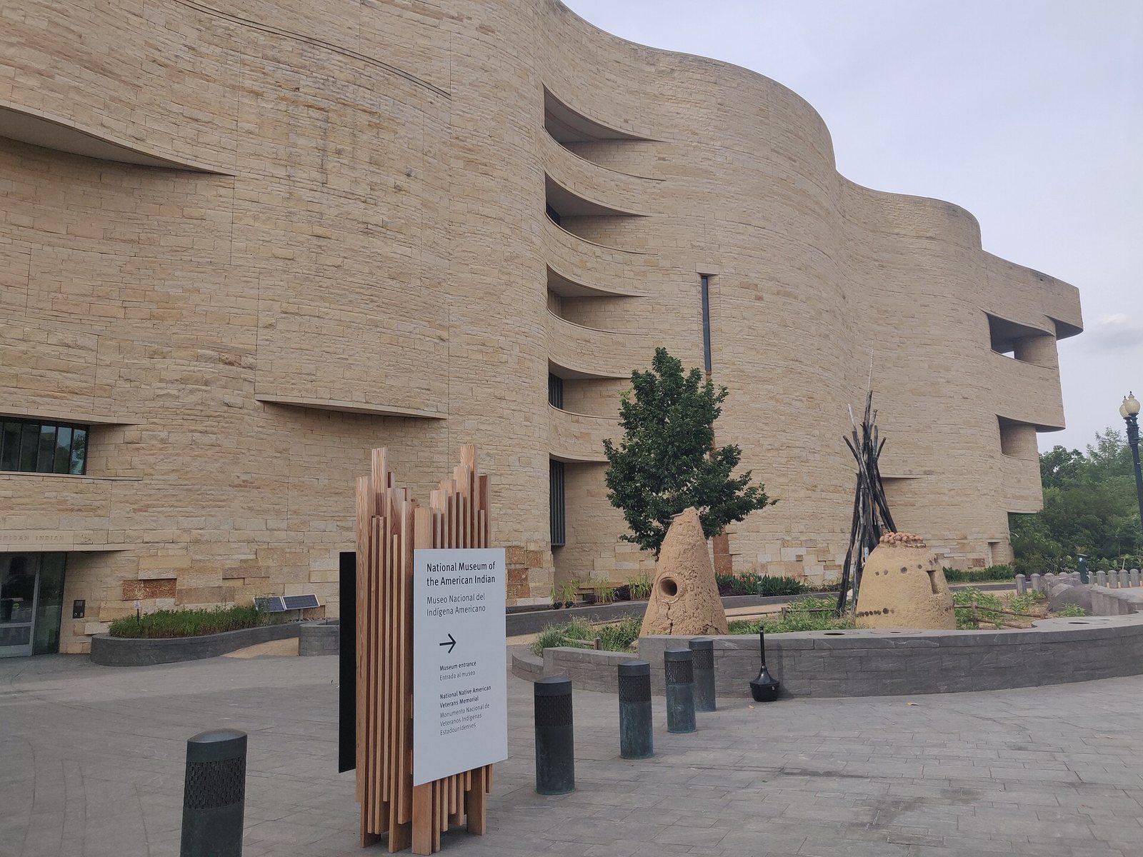 El muro curvo de piedra color miel del Museo Nacional del Indígena Americano en Washington, D. C., con un letrero de madera, un árbol pequeño y dos montículos de tierra con postes en la plaza