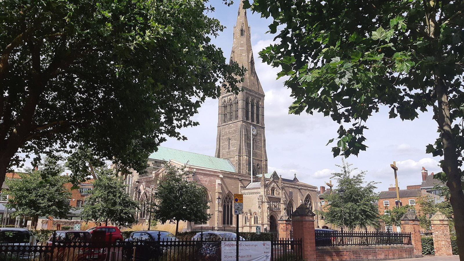Leicester Cathedral with its tall stone spire rising over trees, an iron fence and a street on a bright day
