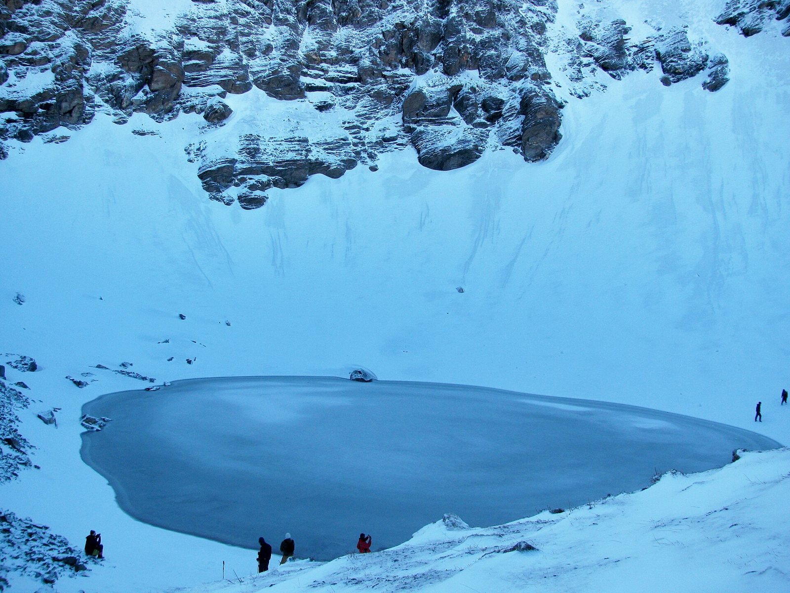 A frozen lake in a steep snow-filled bowl beneath dark cliffs, with a few trekkers standing tiny at the edge