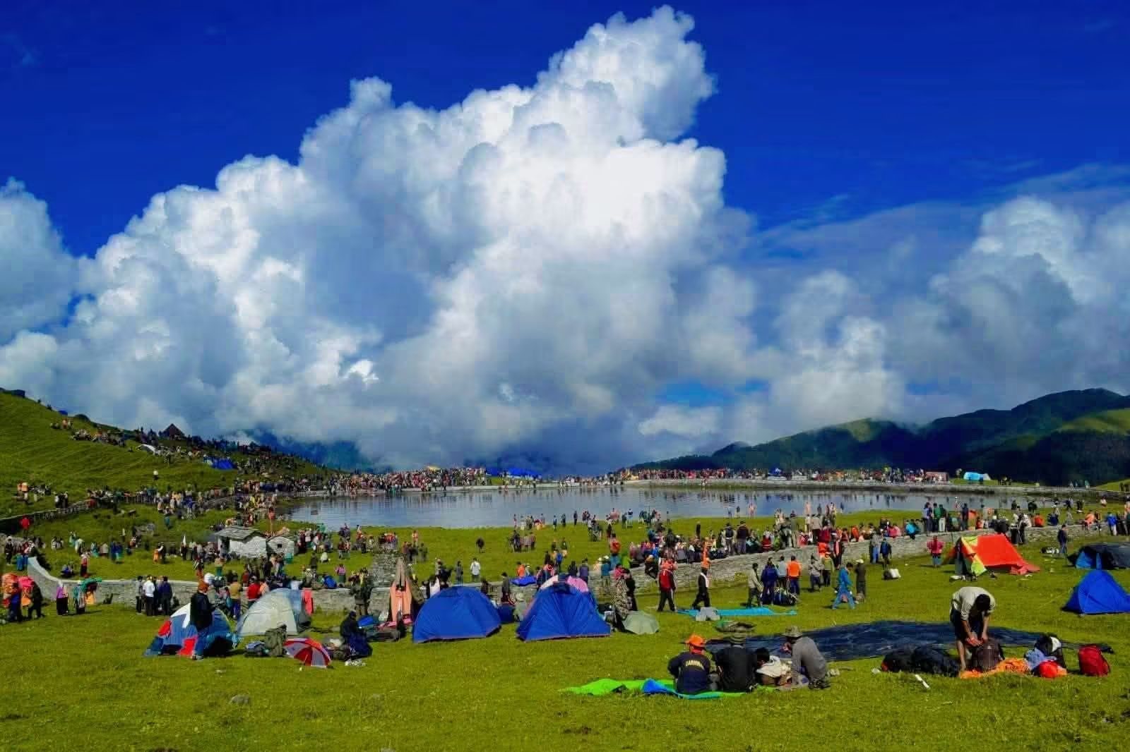 A crowd of pilgrims and many small tents scattered across a green meadow beside a lake, under a huge white cloud in a deep blue sky