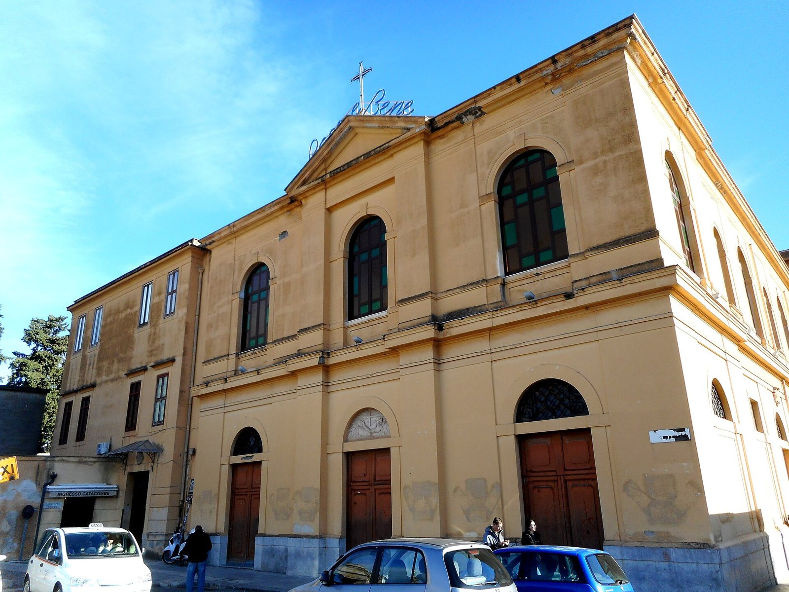 The yellow-ochre front of the church of Santa Maria della Pace dei Cappuccini in Palermo, with arched windows, wooden doors and a cross on the roof, under a blue sky