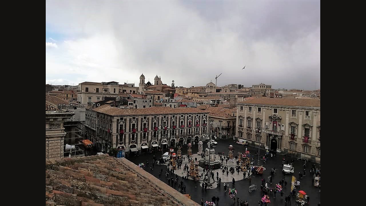 A view from a terrace over a wide square in Catania on a grey day, with crowds below and tall decorated festival structures standing among them during the feast of Saint Agatha