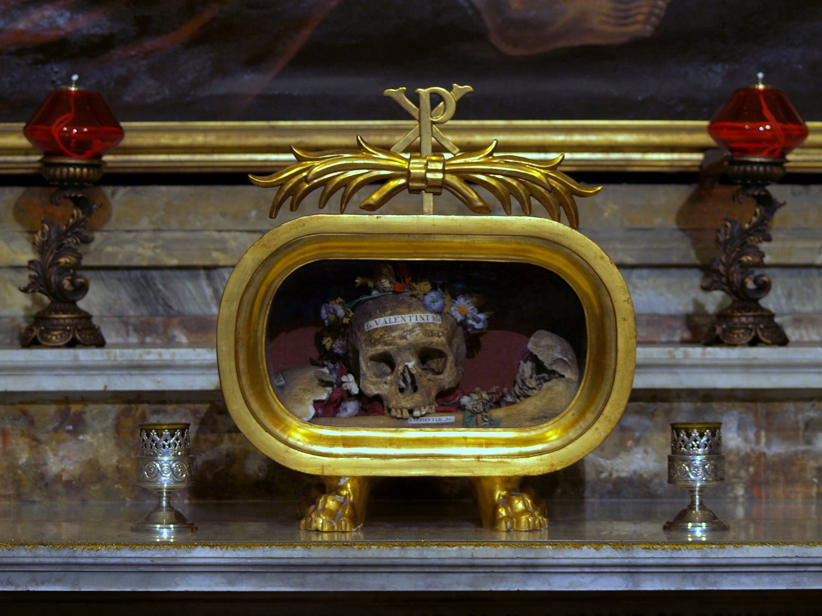 A skull crowned with small flowers inside an oval gilded reliquary topped by a golden Chi Rho, with a name band across the brow, set on a marble altar between two red glass lamps