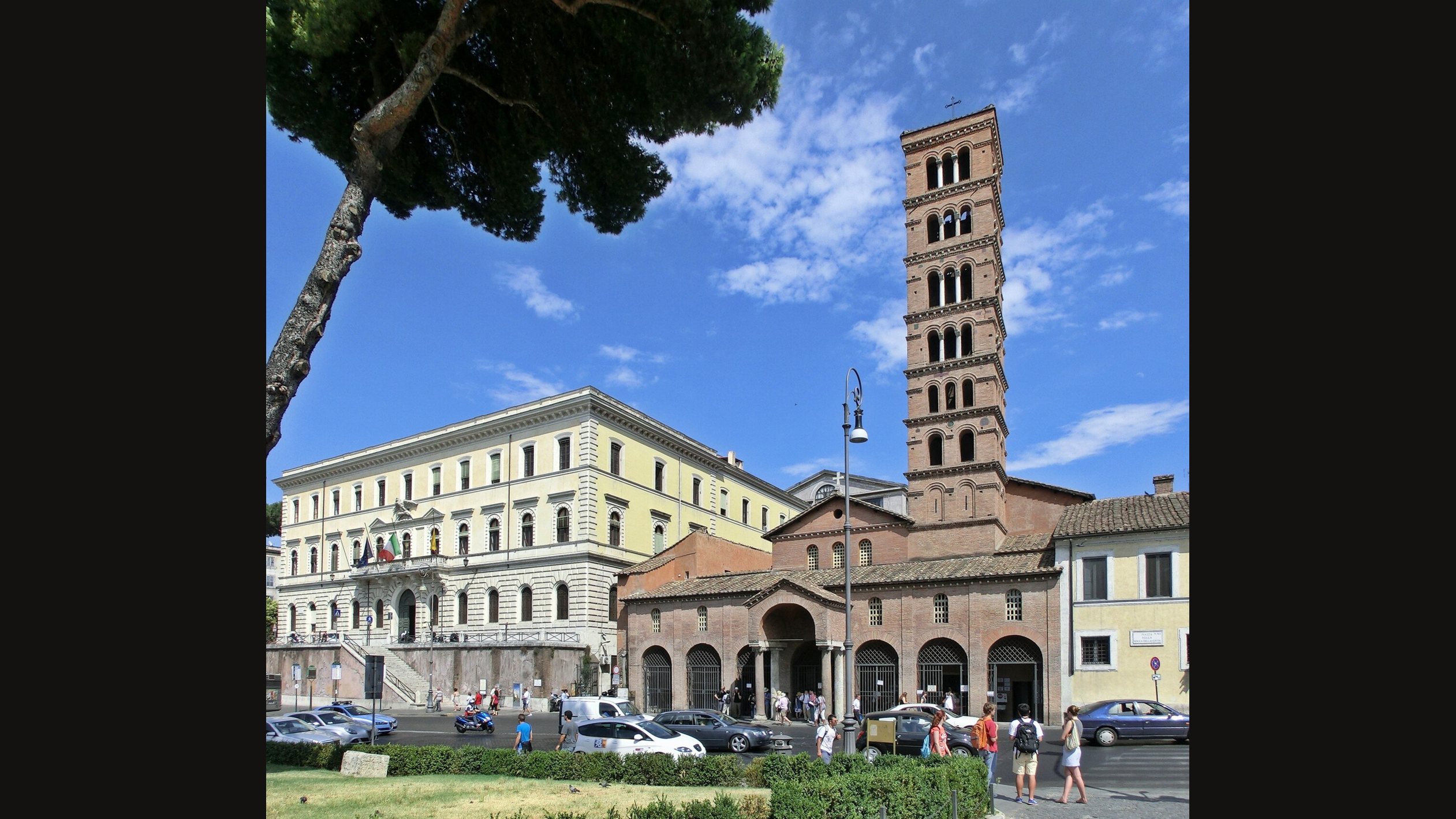 The brick front of Santa Maria in Cosmedin in Rome with its arched portico and tall Romanesque bell tower, beside a pale stone building, seen across a road with parked cars and pedestrians under a blue sky