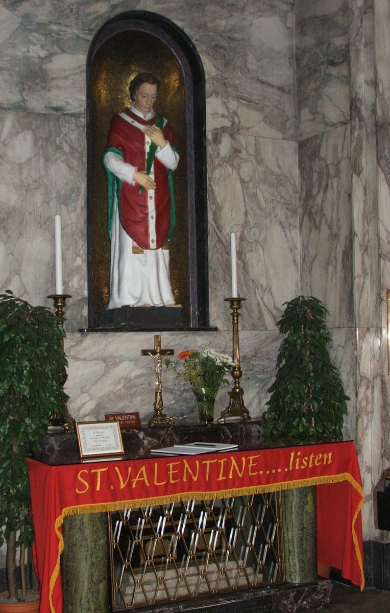 A marble shrine with a painted statue of Saint Valentine in red vestments in an arched niche, above an altar table draped in red cloth reading St. Valentine, with candles, a crucifix and potted plants