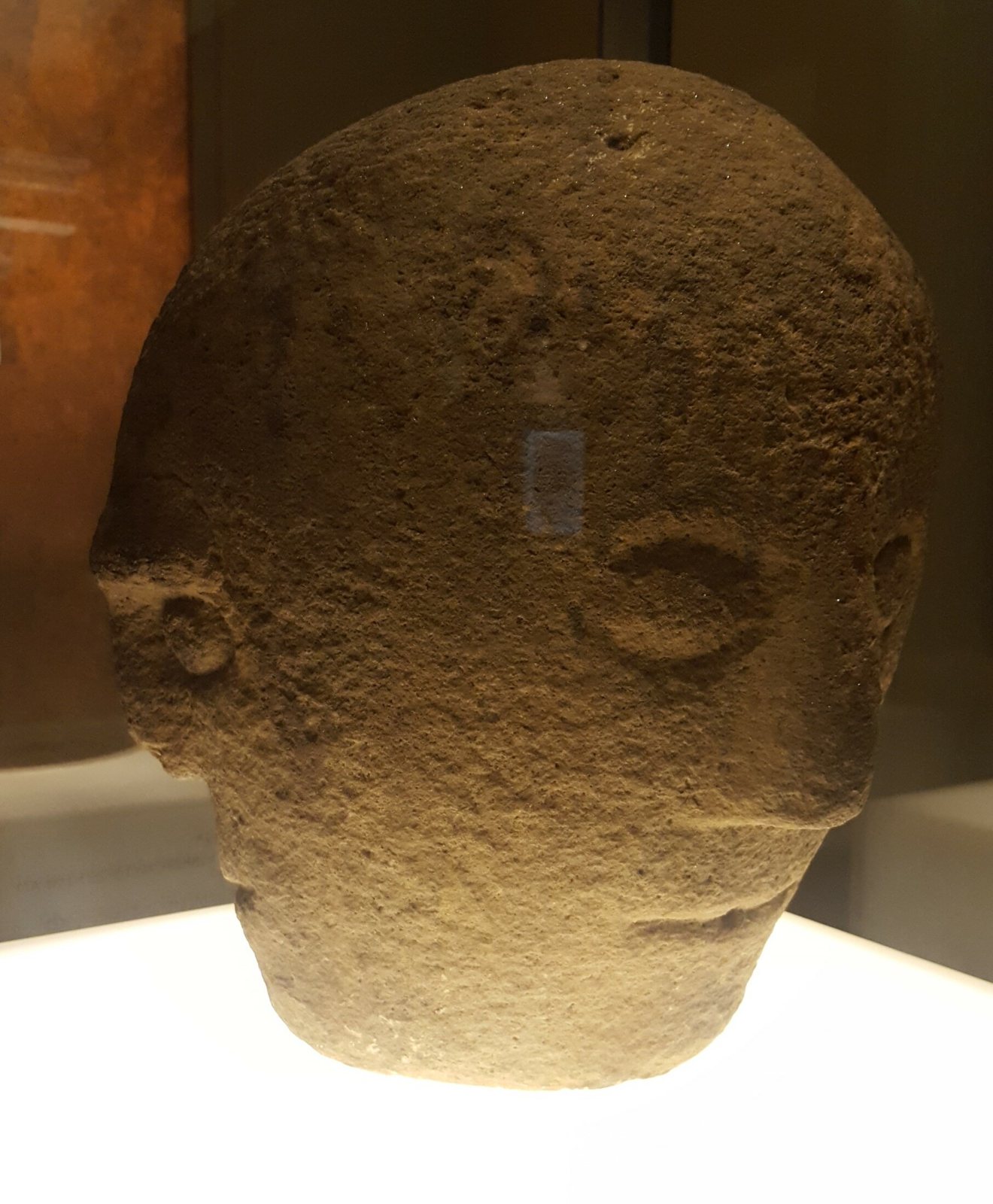 A carved stone head with a smooth domed top, deep-set eyes and a faint nose and mouth, on a pale stand in a museum display