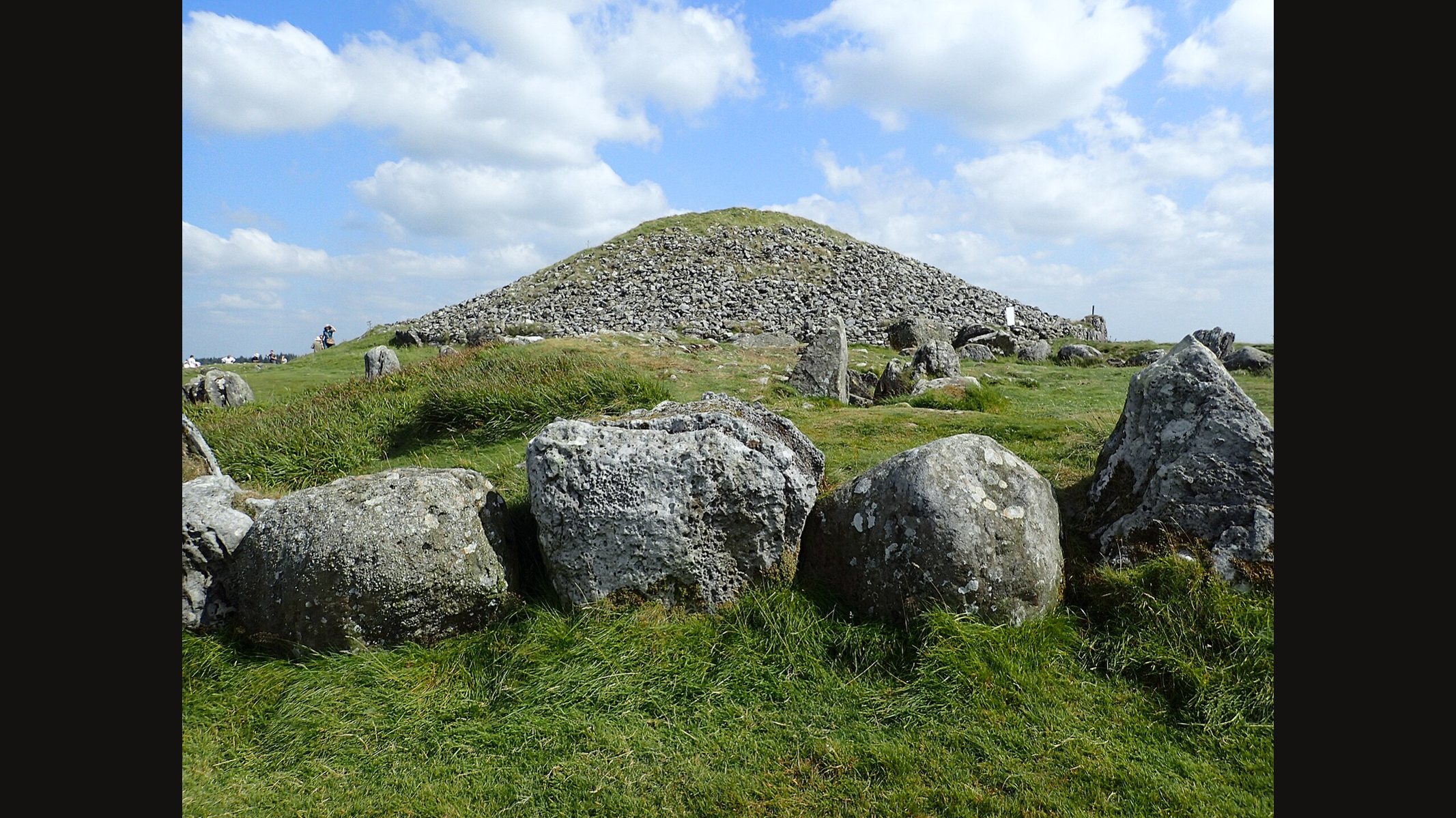Cairn T at Loughcrew in County Meath: a grassy mound capped with grey stones, with large kerbstones in the foreground under a blue sky with white clouds