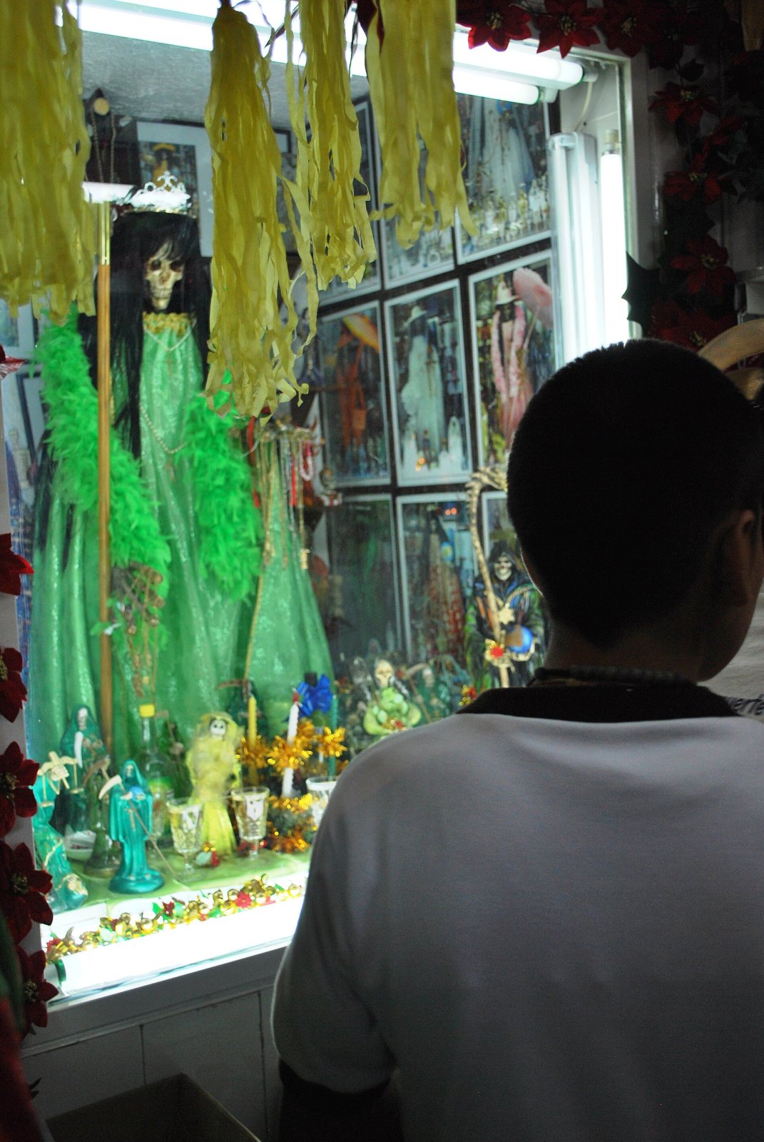 A visitor seen from behind looks through the glass of the Alfarería Street shrine, where a Santa Muerte figure in a long green robe stands among flowers and framed pictures