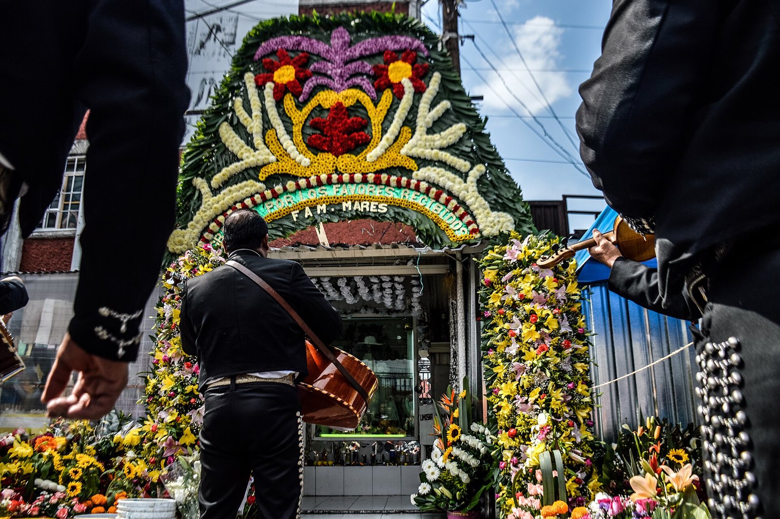 Doña Queta's flower-covered Santa Muerte altar on Alfarería Street in Tepito, seen between two mariachis in black charro suits as a guitarist walks toward it